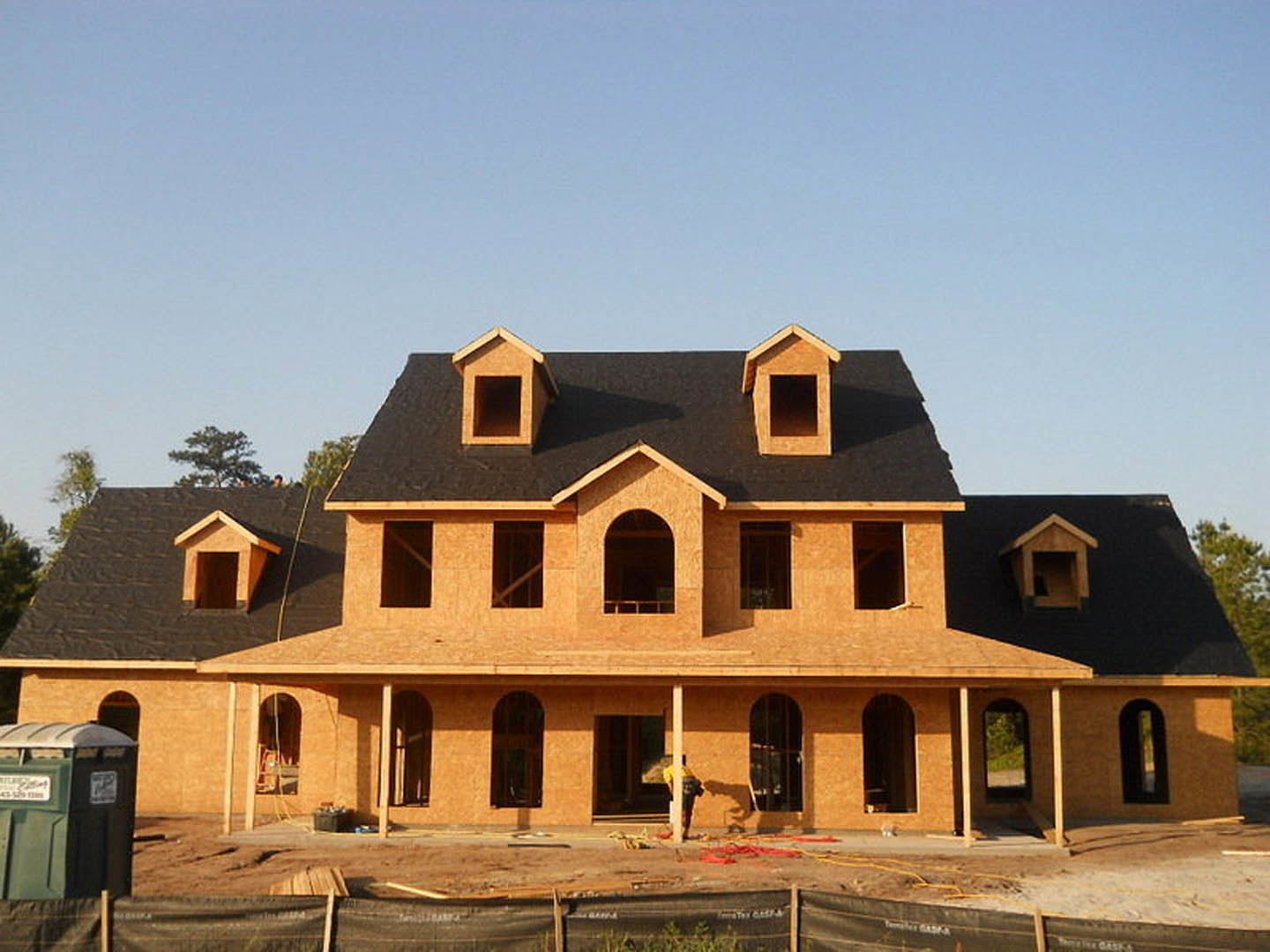 Partially built house with exposed framing, plywood sheathing, and a temporary wooden fence; construction materials scattered in front, man standing near entrance, green trash can