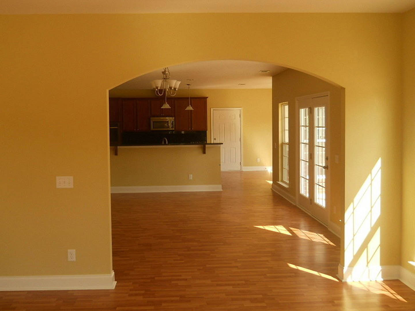 Open-concept kitchen with hardwood floors, white cabinetry, black countertops, and a modern chandelier; white door and large window visible.
