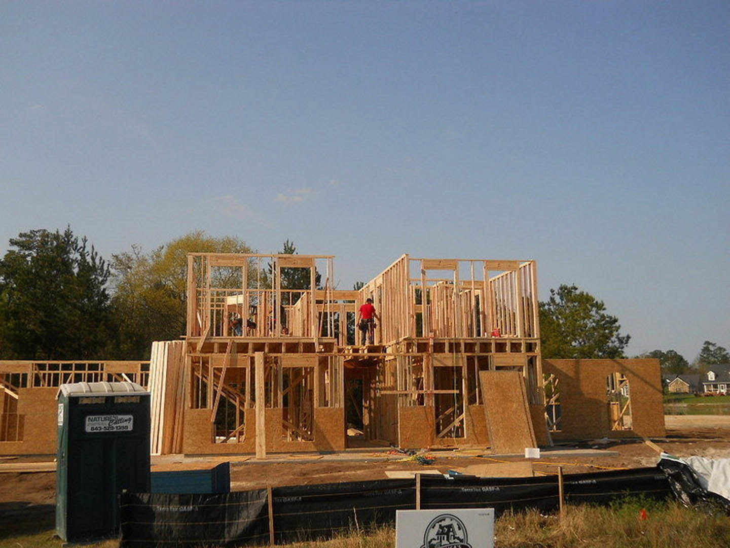 Wood-framed house under construction with workers on site, exposed lumber, partially completed exterior, blue sky, and surrounding trees