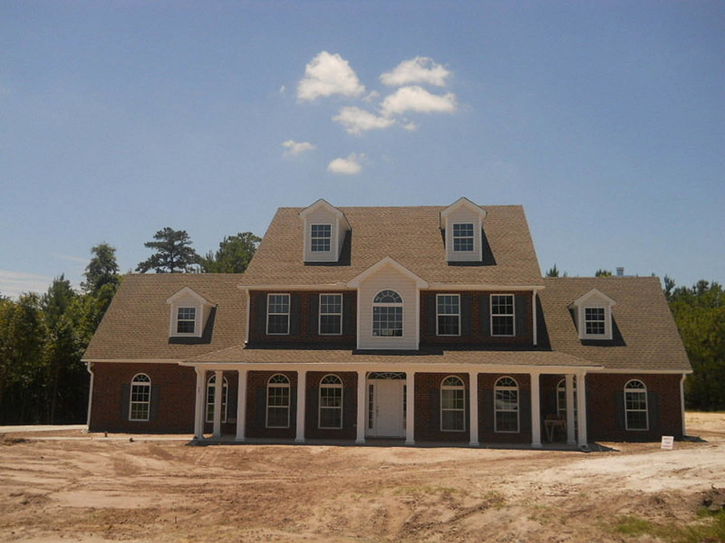 Spacious two-story home under construction with white door featuring glass panel, white-framed windows, unfinished exterior, dirt yard, and partly cloudy sky