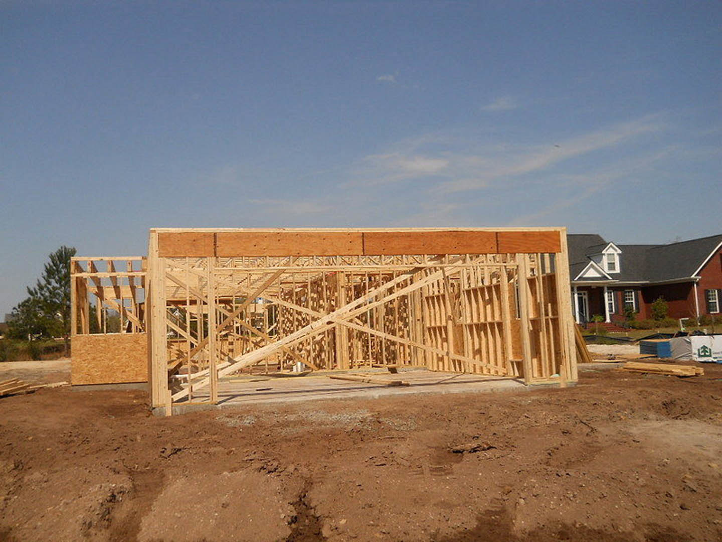 Wooden framing of a house under construction on a dirt lot, with a finished home featuring a white roof visible in the background, surrounded by trees and construction materials.