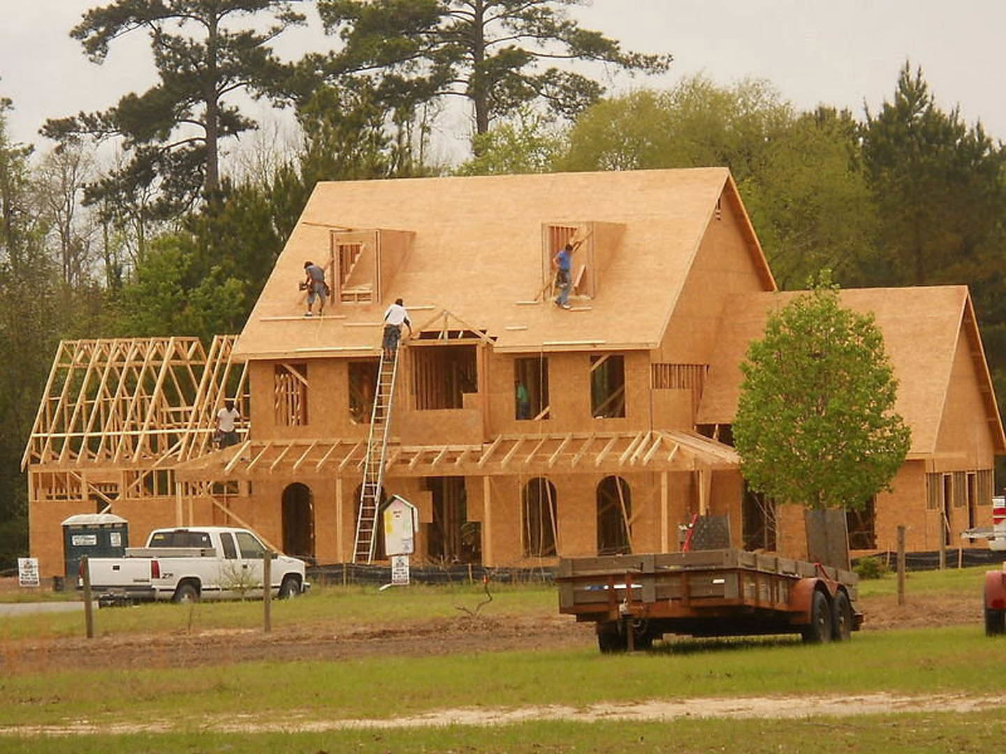 Framed house under construction with several workers, white truck parked on grassy field, leafy tree nearby, blue sky overhead