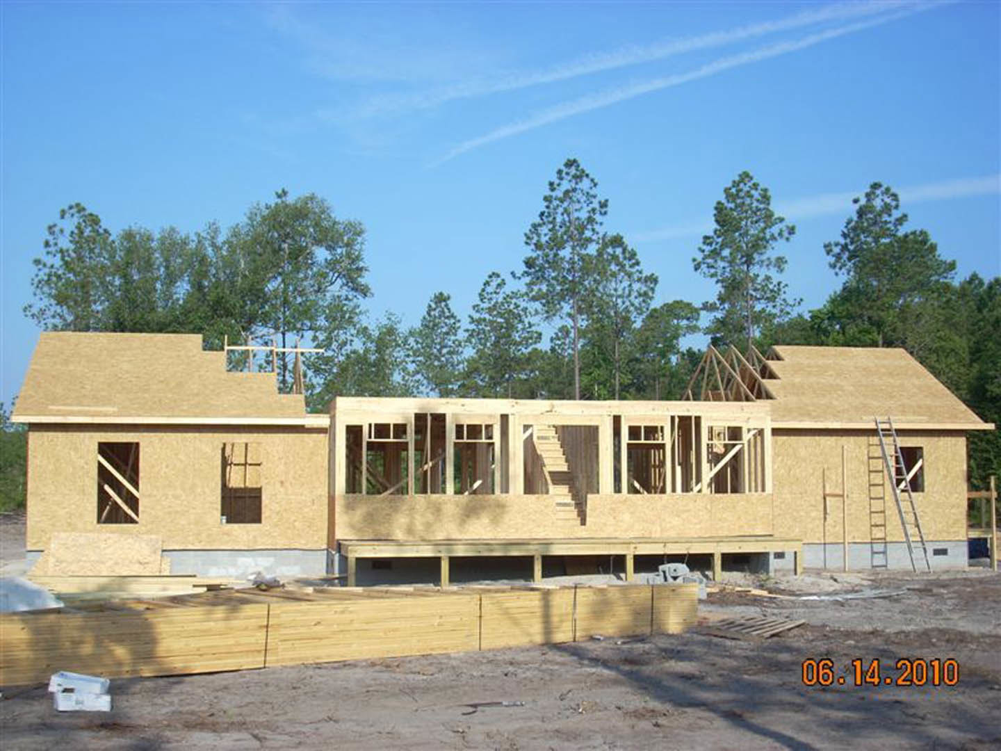 Framed house under construction with exposed wooden beams, unfinished staircase, stacked lumber on dirt, large windows, and leafy trees in the background