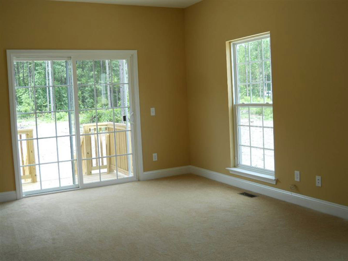 Beige carpeted room featuring a sliding glass door with wooden railing, large windows offering views of trees, and white walls with natural daylighting.
