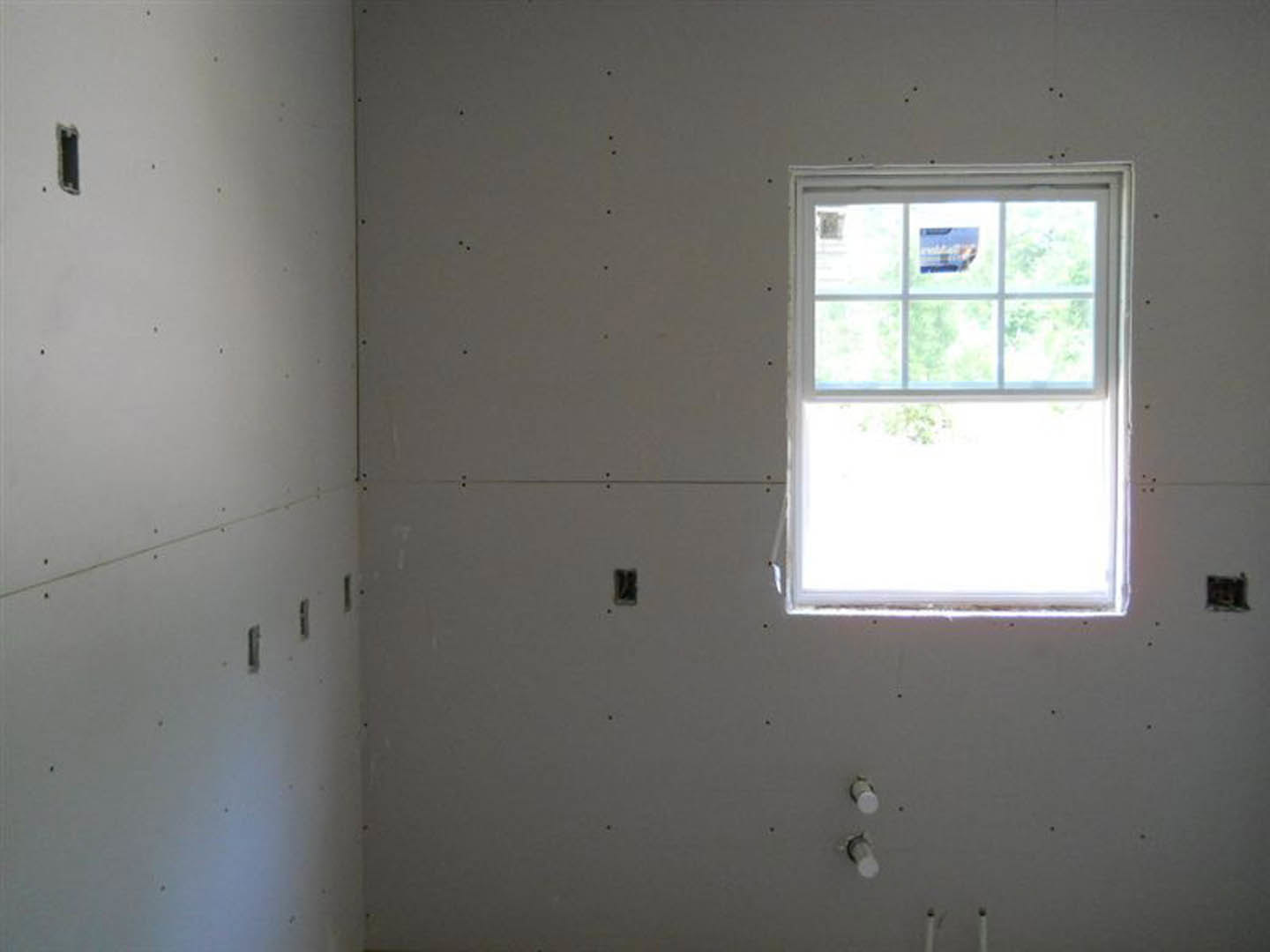 White drywall wall with several holes, window with sticker, light switches, blue sky visible through glass