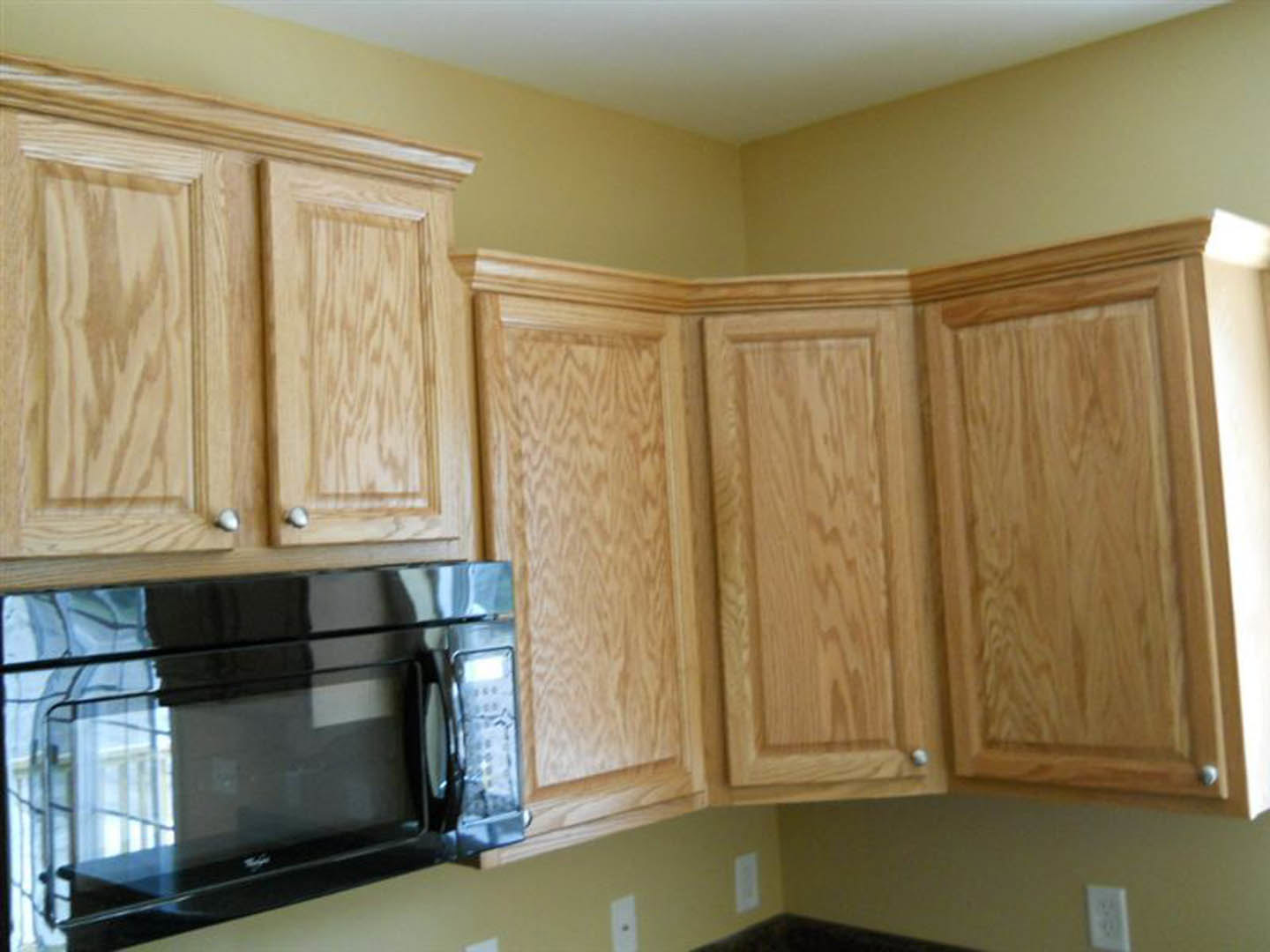 Kitchen with natural wood cabinets, black microwave oven, white electrical outlet, and light-colored countertop