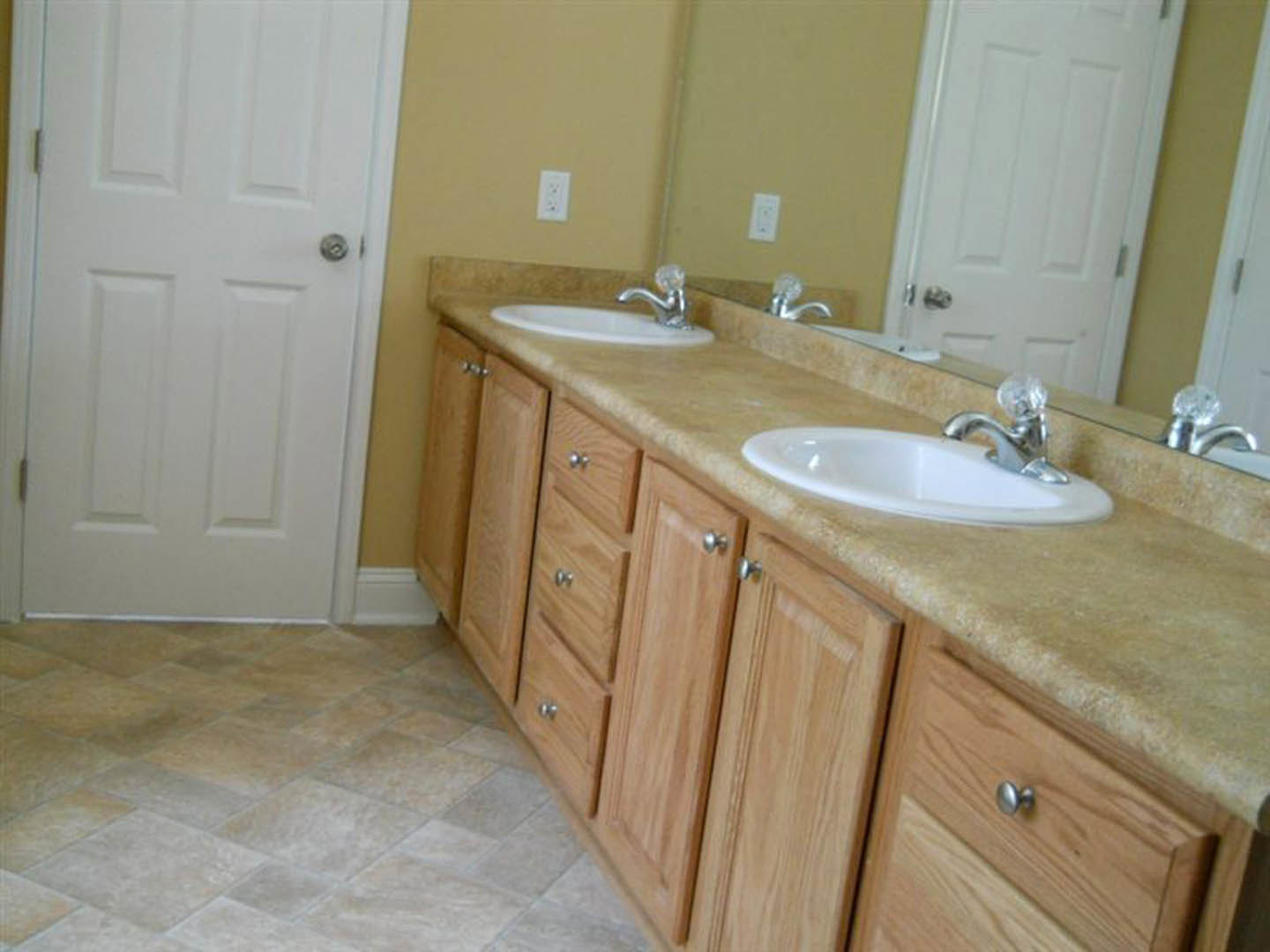 Double vanity with white sinks and silver faucets, wood cabinetry, tile flooring, white door with silver handle, bathroom accessories