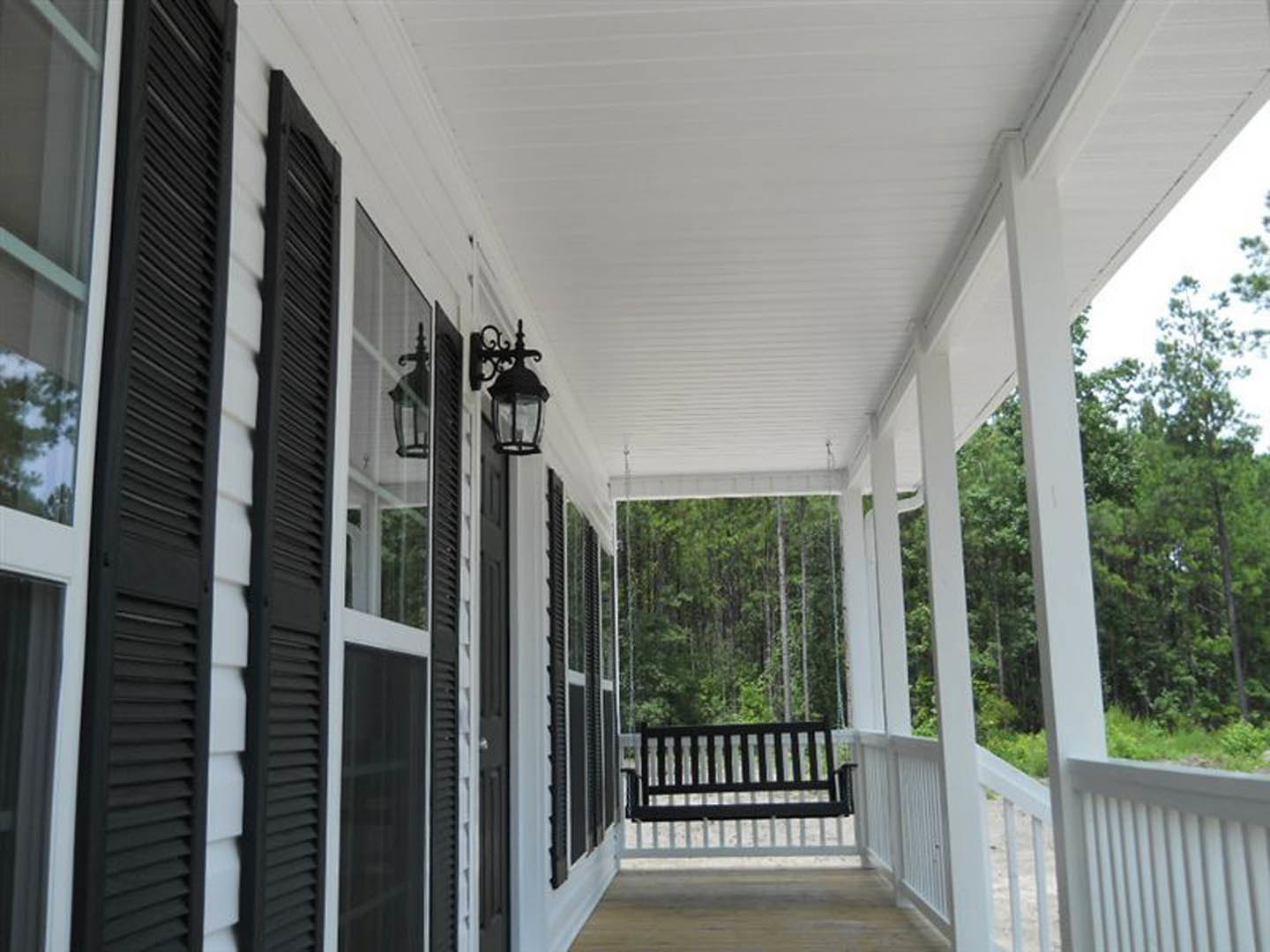 White porch with square pillars, wooden swing, and bench; black light fixture mounted on white siding; tree branches visible in foreground.