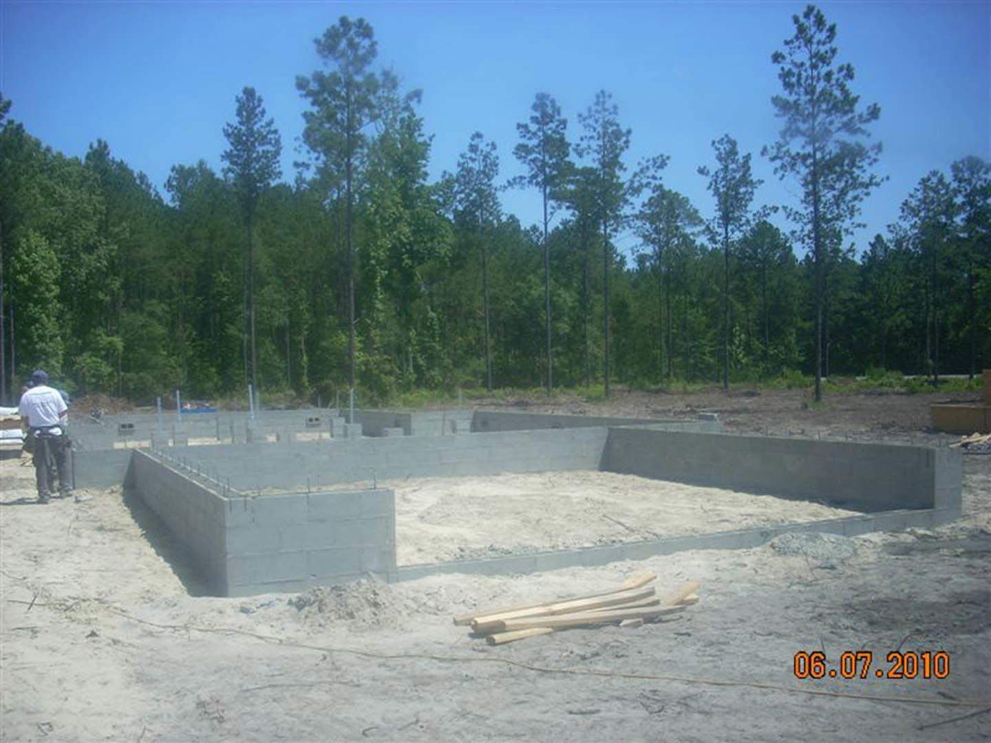 Concrete foundation of a house under construction surrounded by snow, white construction materials, and forest trees; man in white shirt and black pants standing nearby.