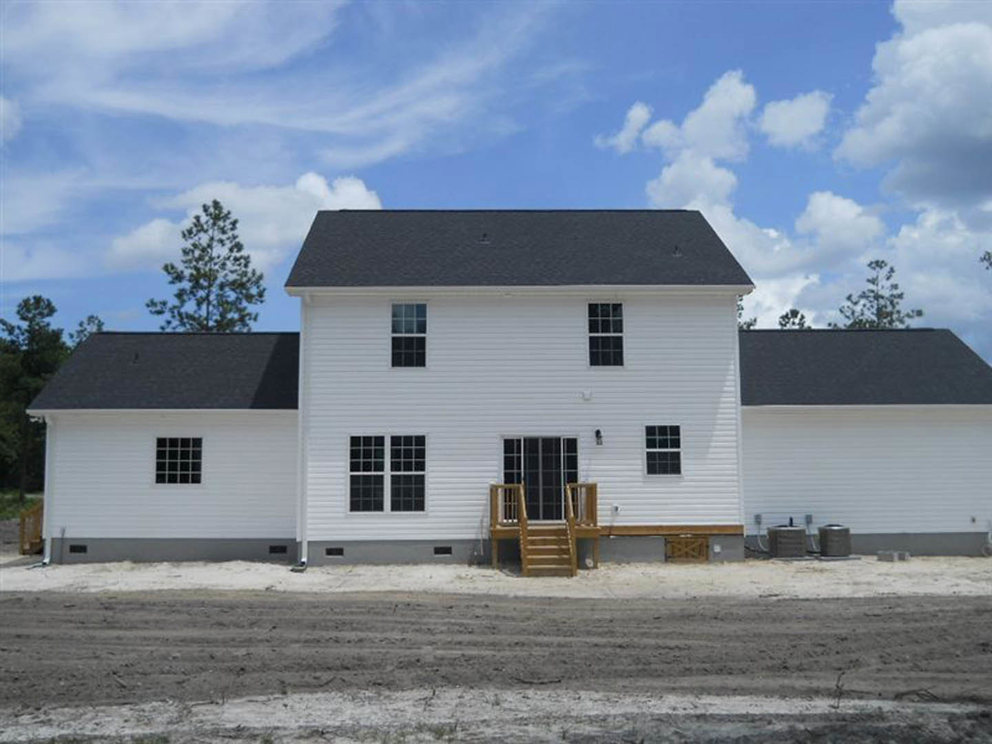 White siding house with black shingle roof, wooden staircase leading to entry, dirt yard in foreground, blue sky with scattered clouds overhead