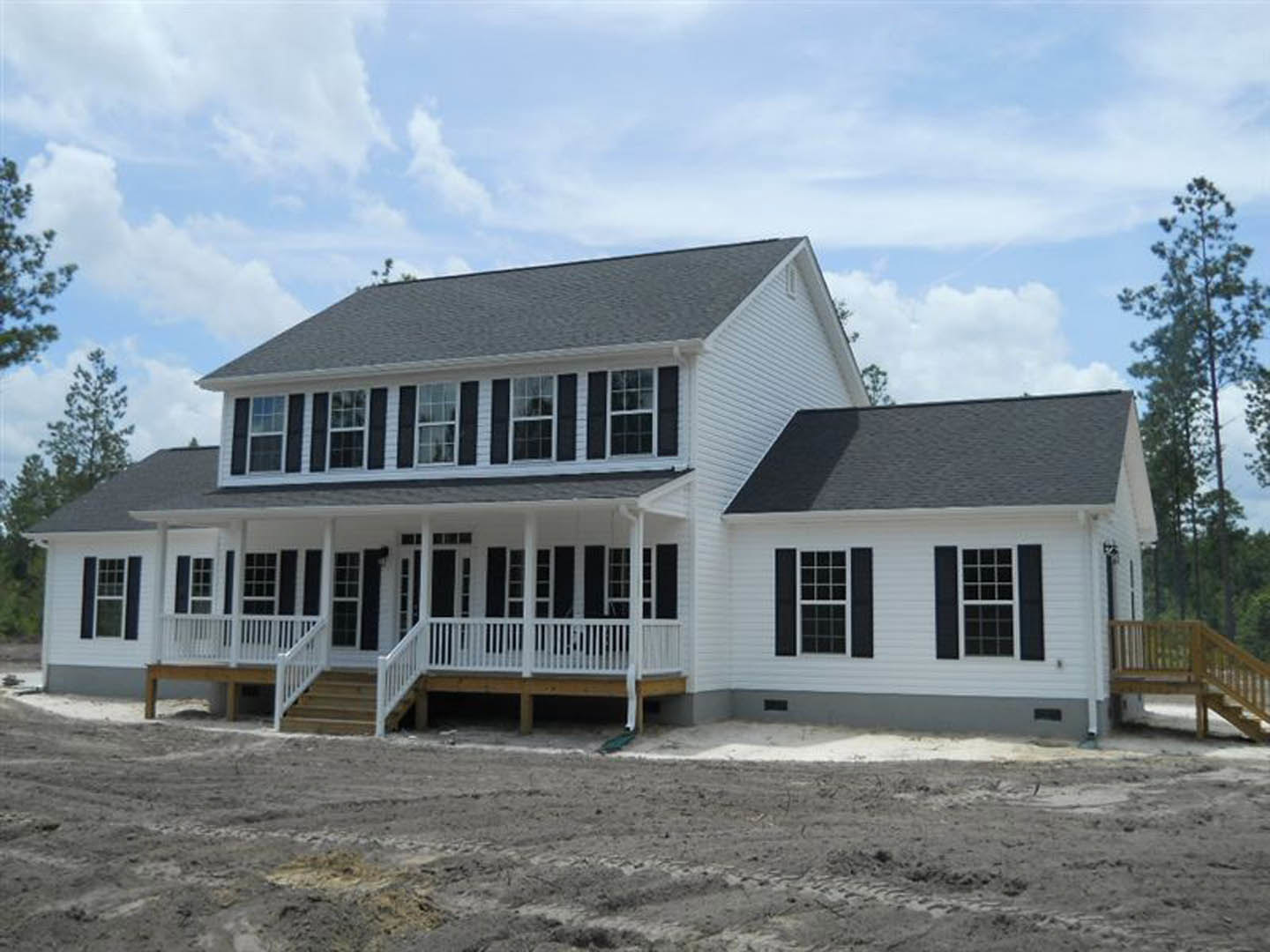 White house with spacious covered porch, wooden railing, multiple windows, tree and open dirt field in background under partly cloudy sky