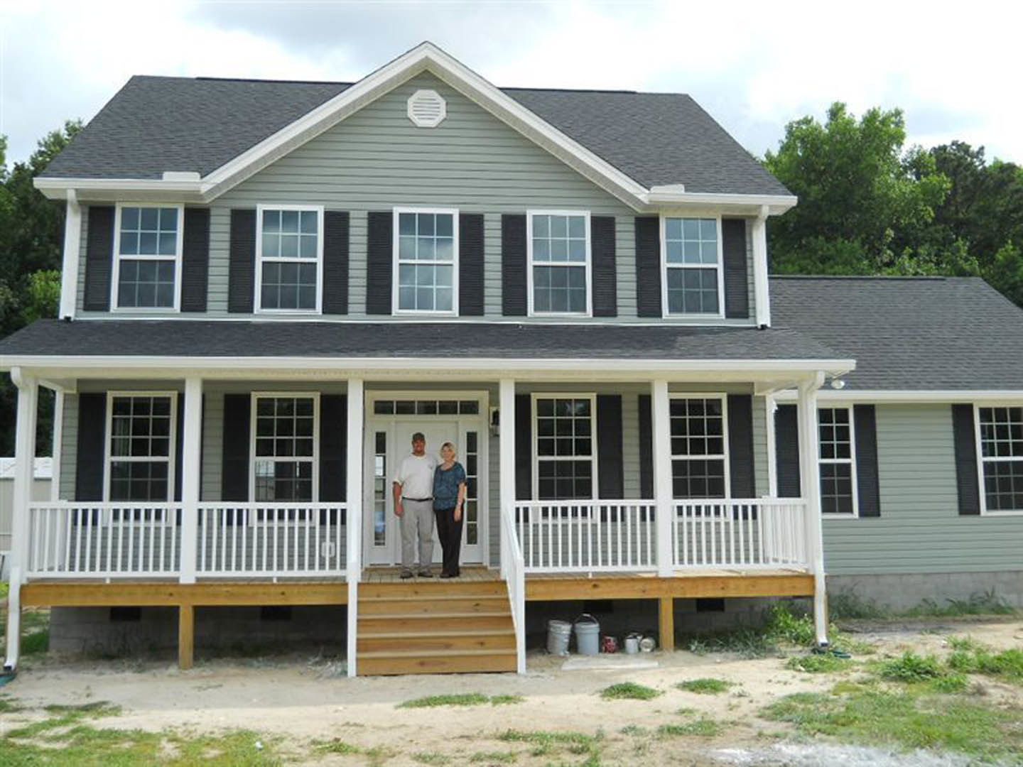 Couple standing on covered porch of grey-sided house with white trim and windows, wooden steps leading up to entry