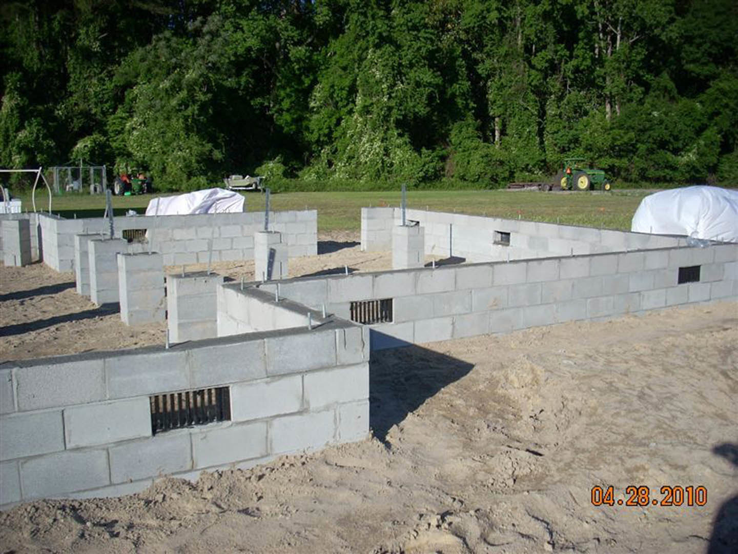 Concrete block foundation with stone accents, green tree with white flowers nearby, grey block wall featuring a vent, white sheet draped over a vehicle, tractor close-up in