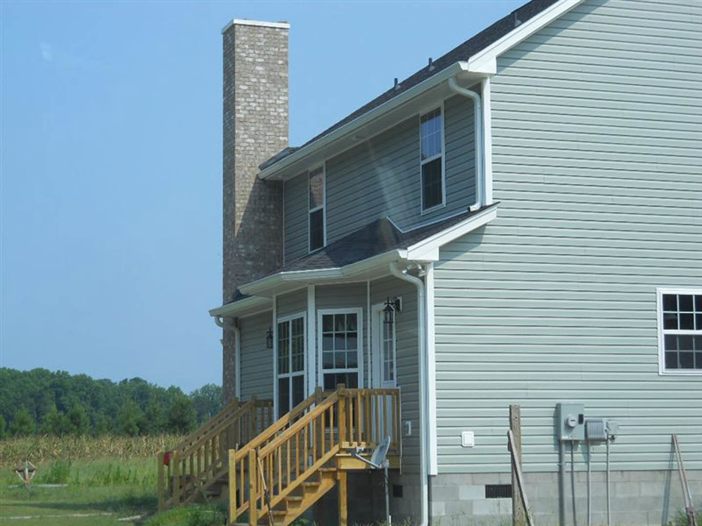 Two-story home with white siding, brick chimney, multi-pane windows, and wooden exterior staircase, surrounded by grass and mature trees.