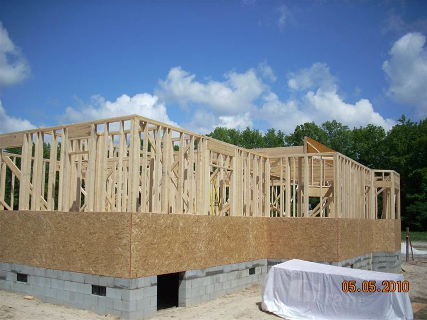 Partially constructed residential building wrapped in white protective sheeting, exposed concrete walls, construction materials on ground, blue sky with scattered clouds overhead