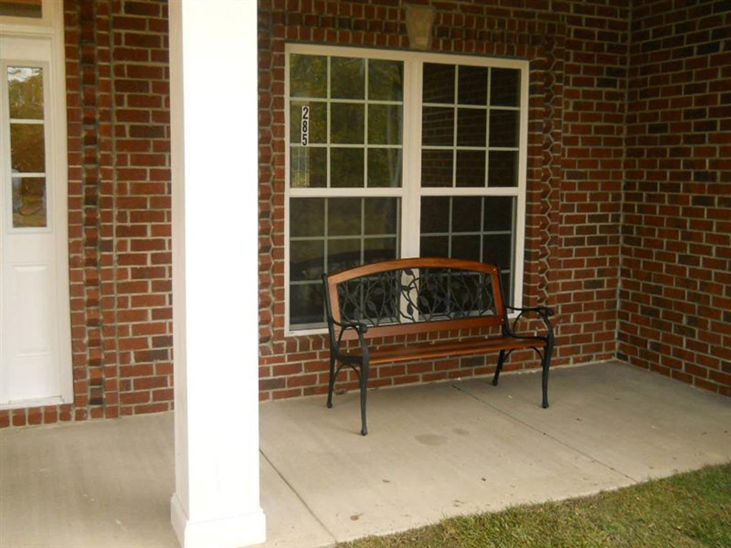 Wooden bench on brick porch beside white door with glass panes, outdoor seating area with grass visible in foreground
