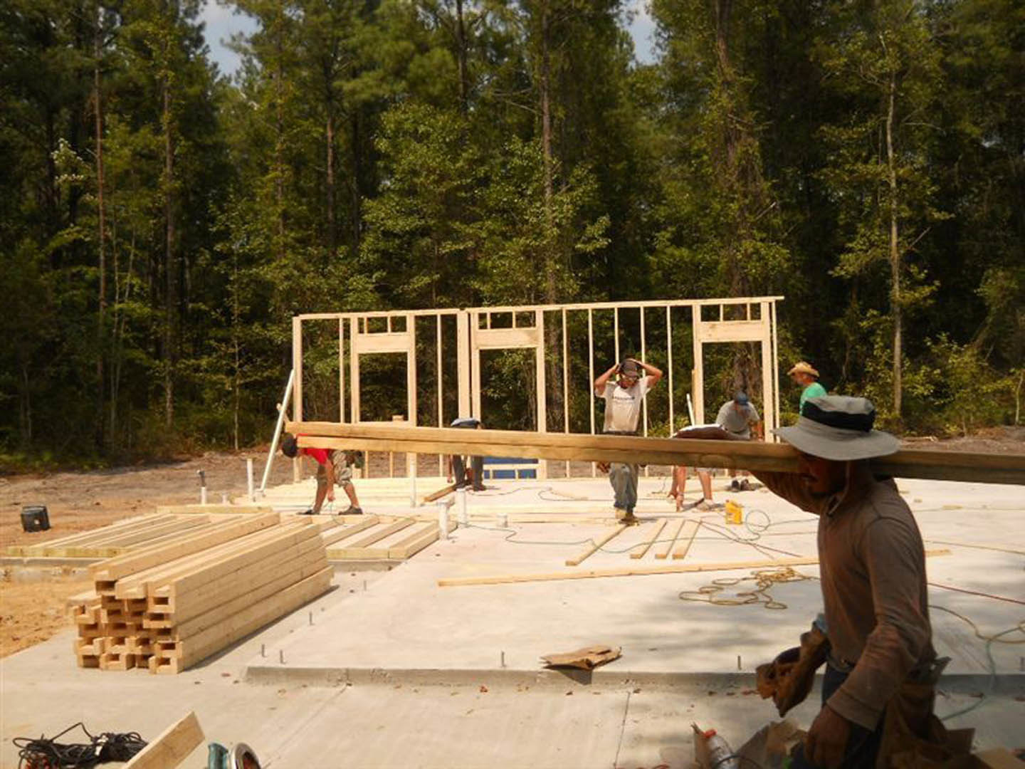 Framed wooden house under construction with several people working, stack of wood beams in foreground, white sign on pole, trees and fencing in background