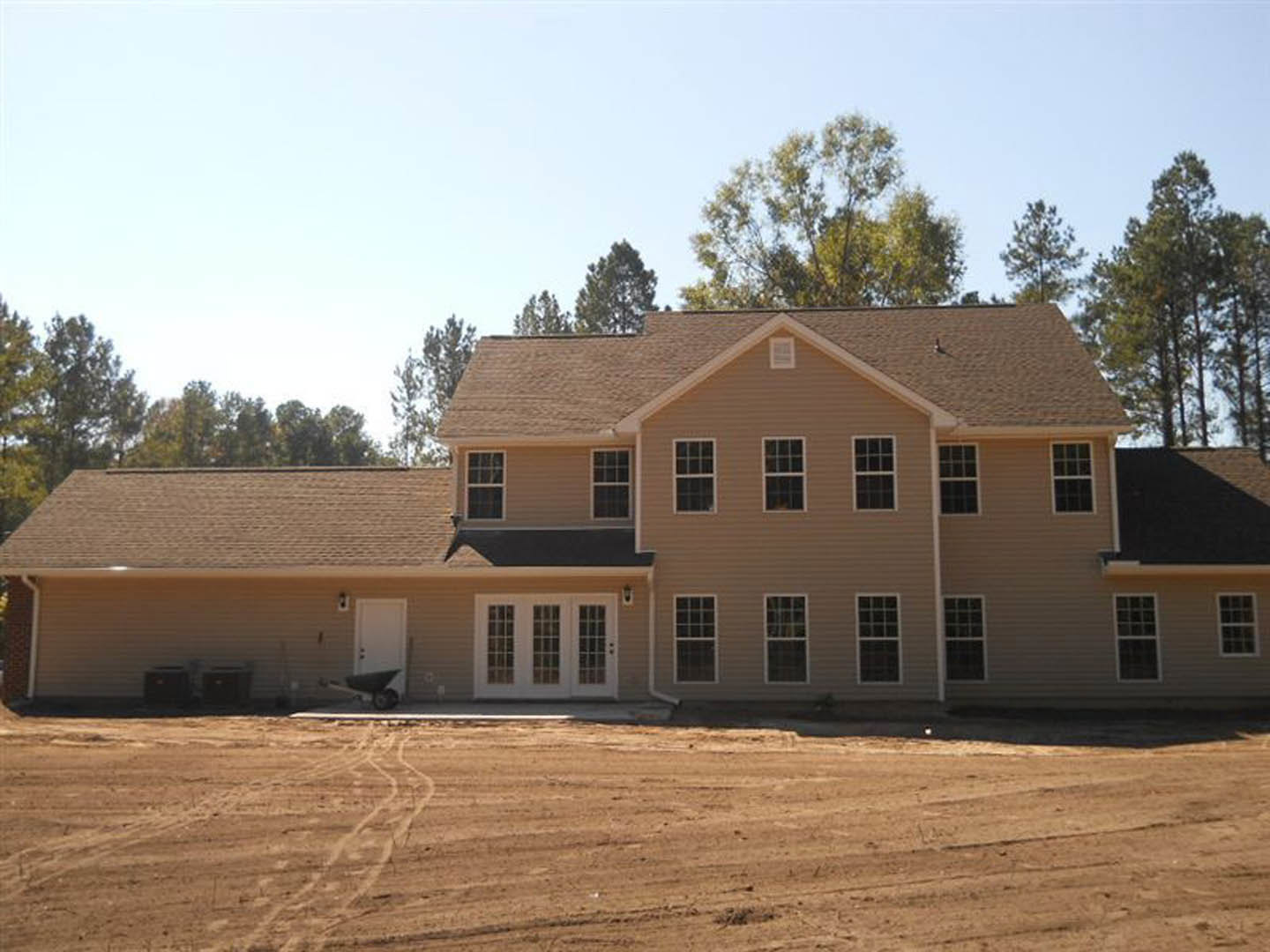 Two-story house with gray roof, white siding, attached garage, multiple windows, and unfinished dirt yard in front, surrounded by trees under blue sky