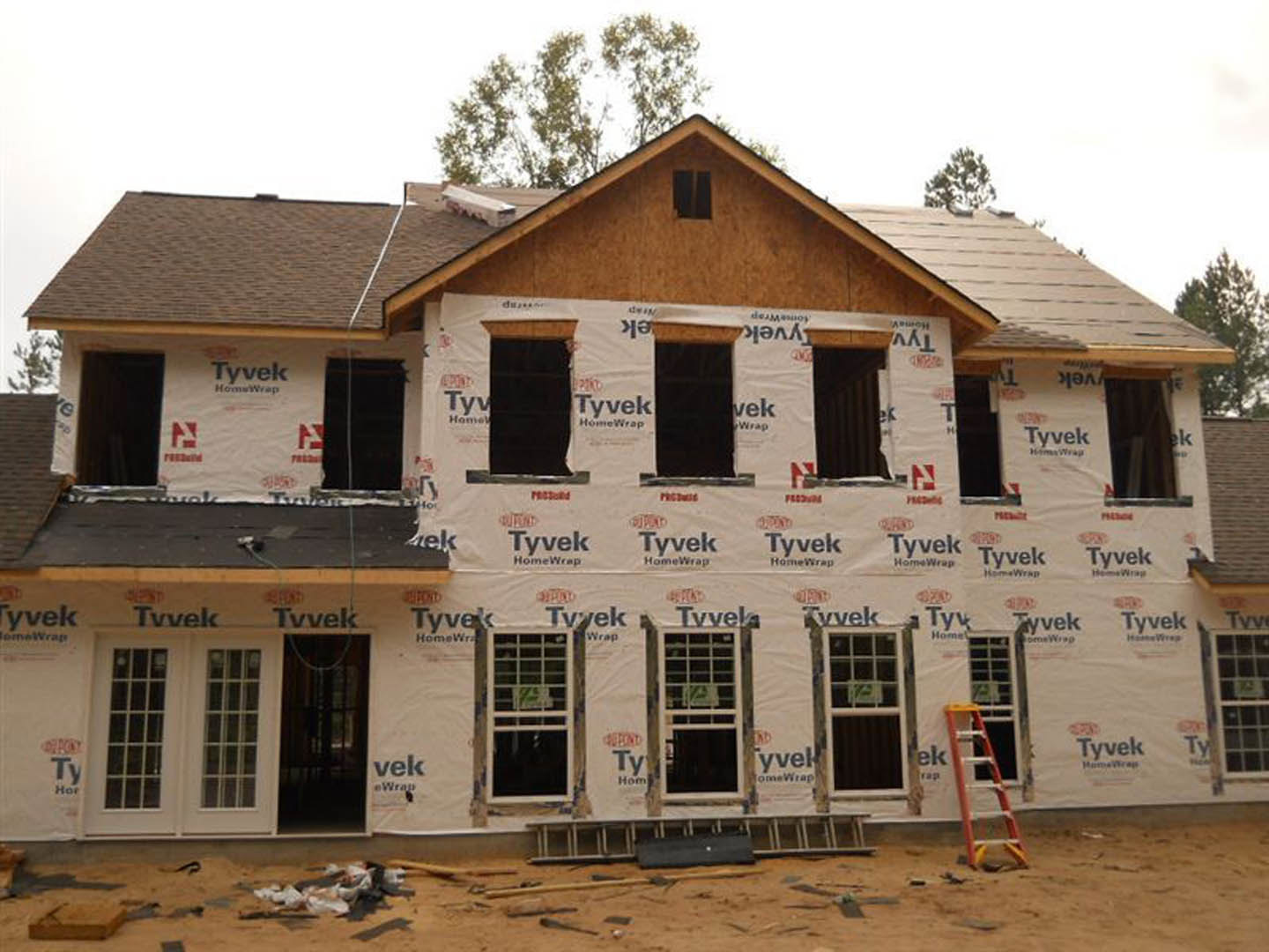 Modern home under construction featuring white exterior walls, black-framed windows with signage, aluminum ladder leaning against the facade, and a pile of tools scattered on the