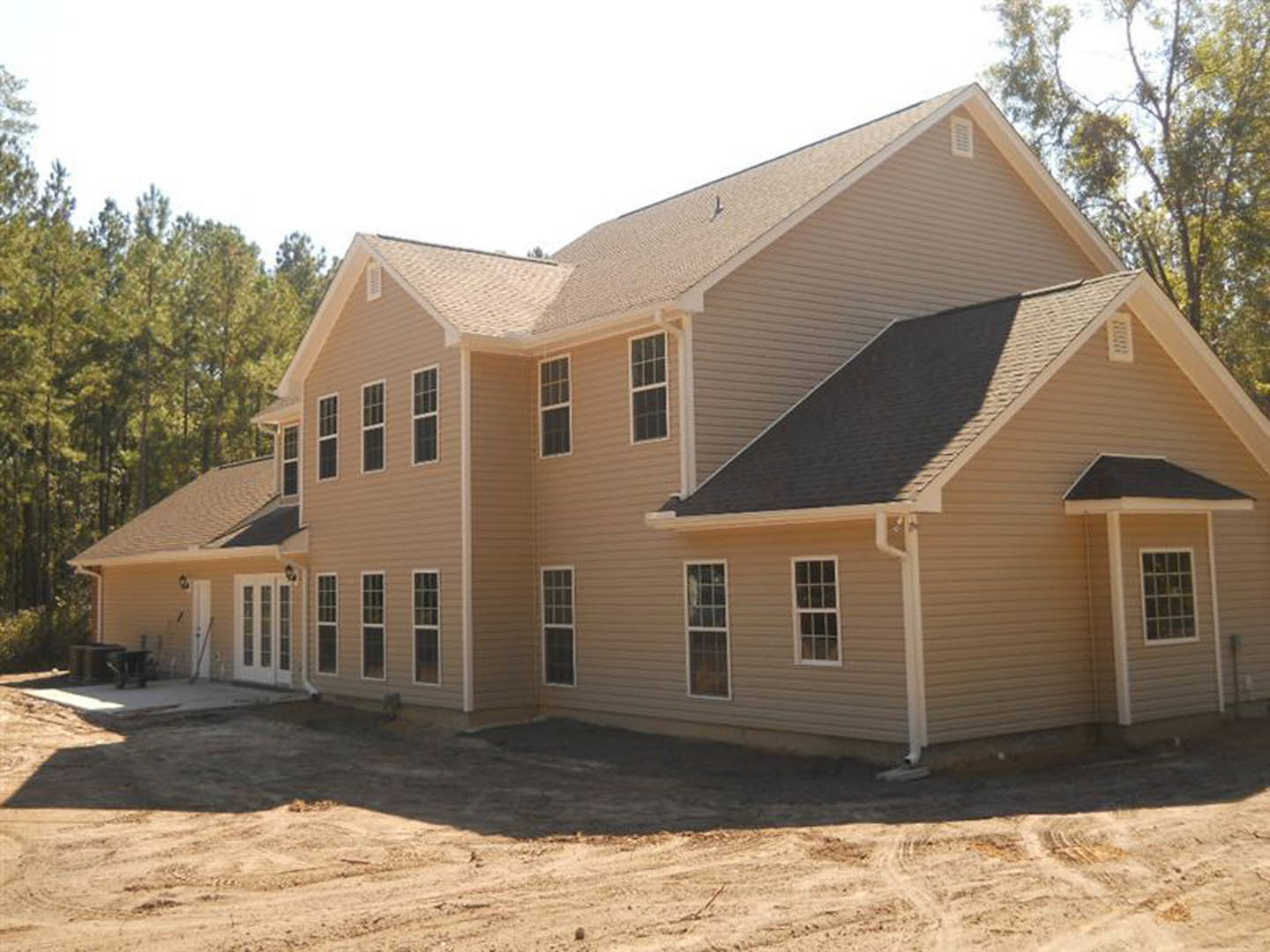 Large tan house with white roof, multiple white-framed windows, paved driveway, dirt patch near entrance, surrounded by trees under blue sky