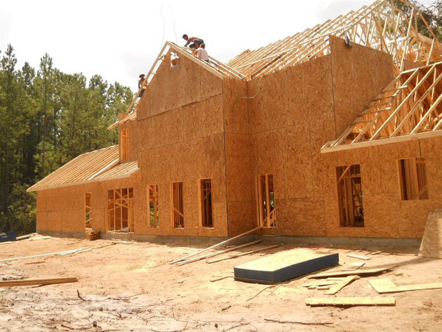 Wood-framed house under construction with exposed beams, workers on site, ladder in doorway, brick exterior partially installed, trees and sky in background