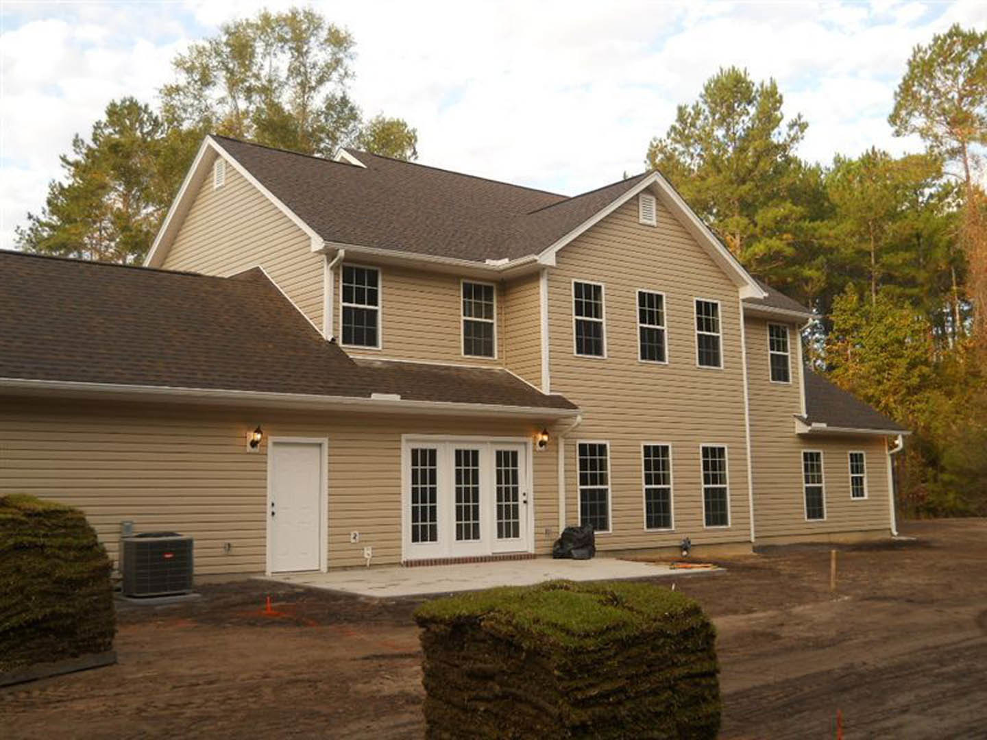 Modern home with white siding, double glass doors, black hardware, surrounded by dirt, grass, and mature trees; driveway and outdoor air conditioning unit visible.