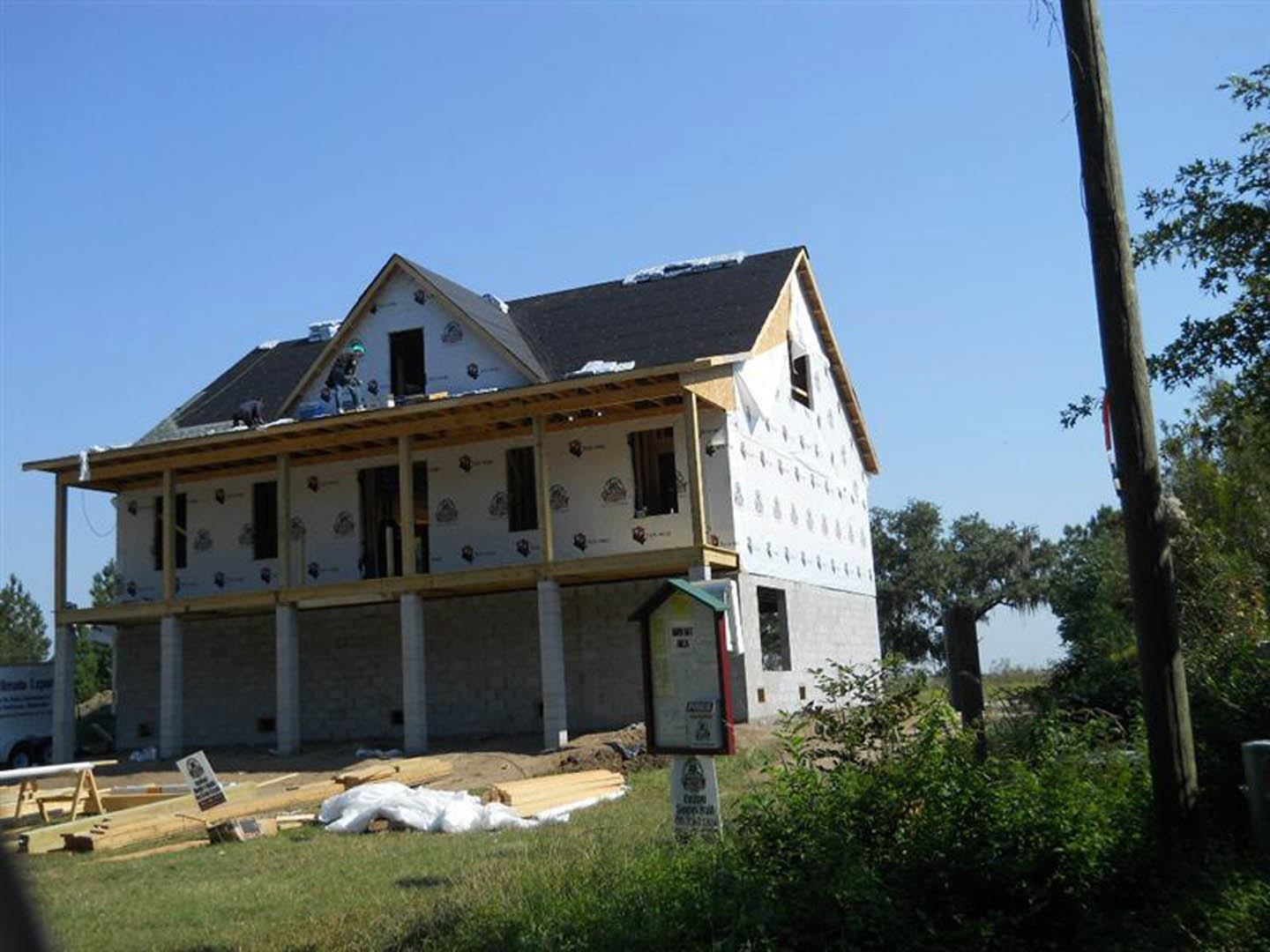 Framed house under construction with exposed wood beams, workers on roof, American flag hanging, tree trunk with red ribbon in foreground, partially visible porch and windows