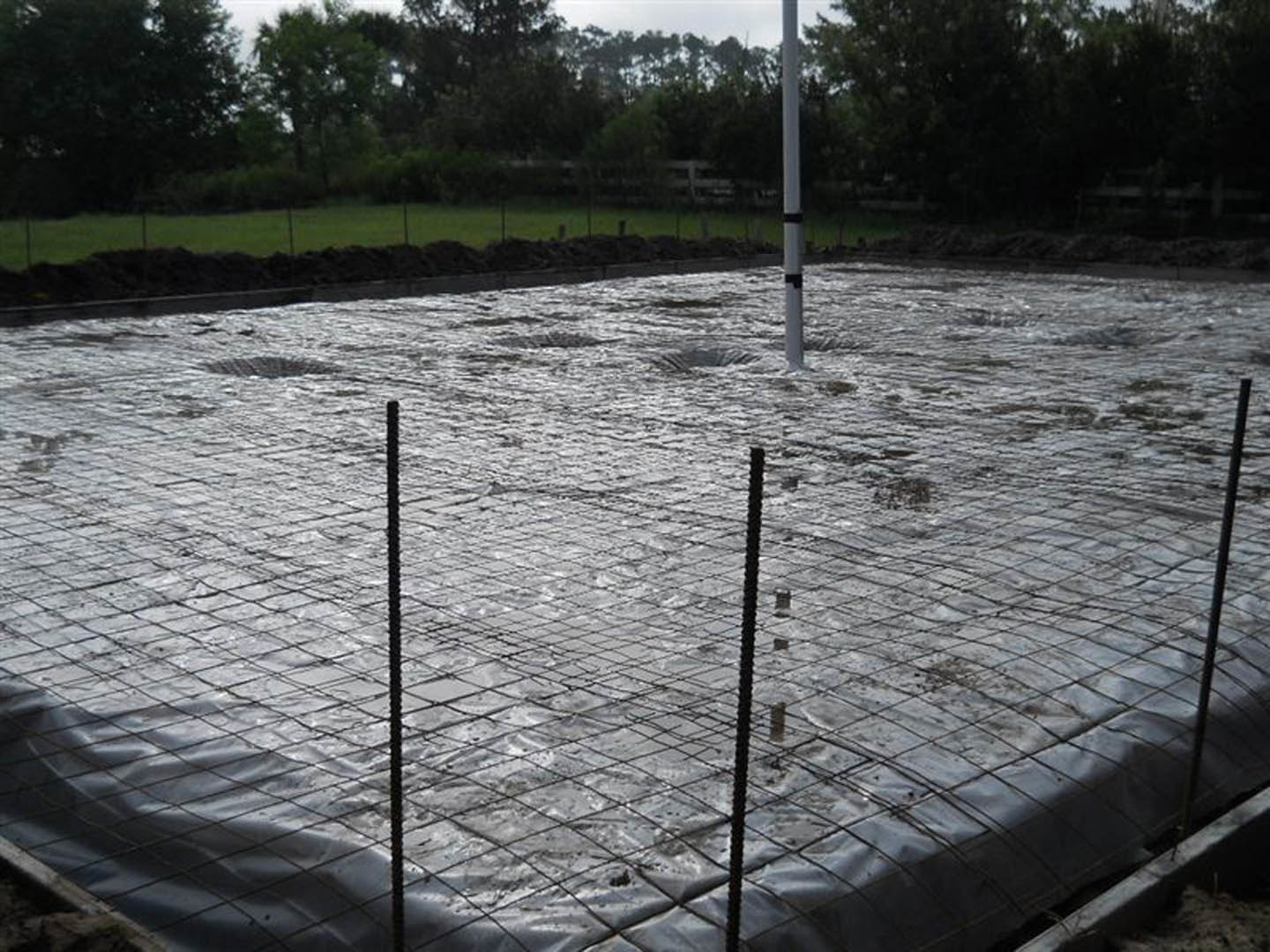 Wet concrete foundation with embedded metal rods and wire mesh, surrounded by construction poles; distant trees visible beyond the site.