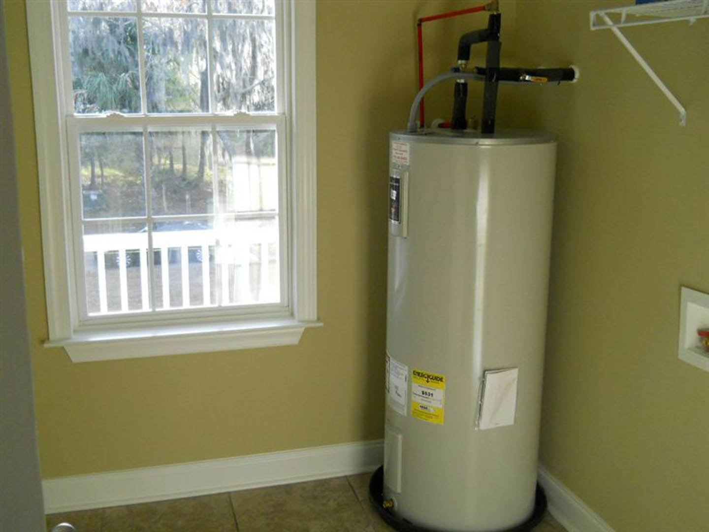 White cylindrical fire extinguisher with black handle and yellow label mounted on wall near window with tree view, white metal bracket, and close-up of pipe and cloth.