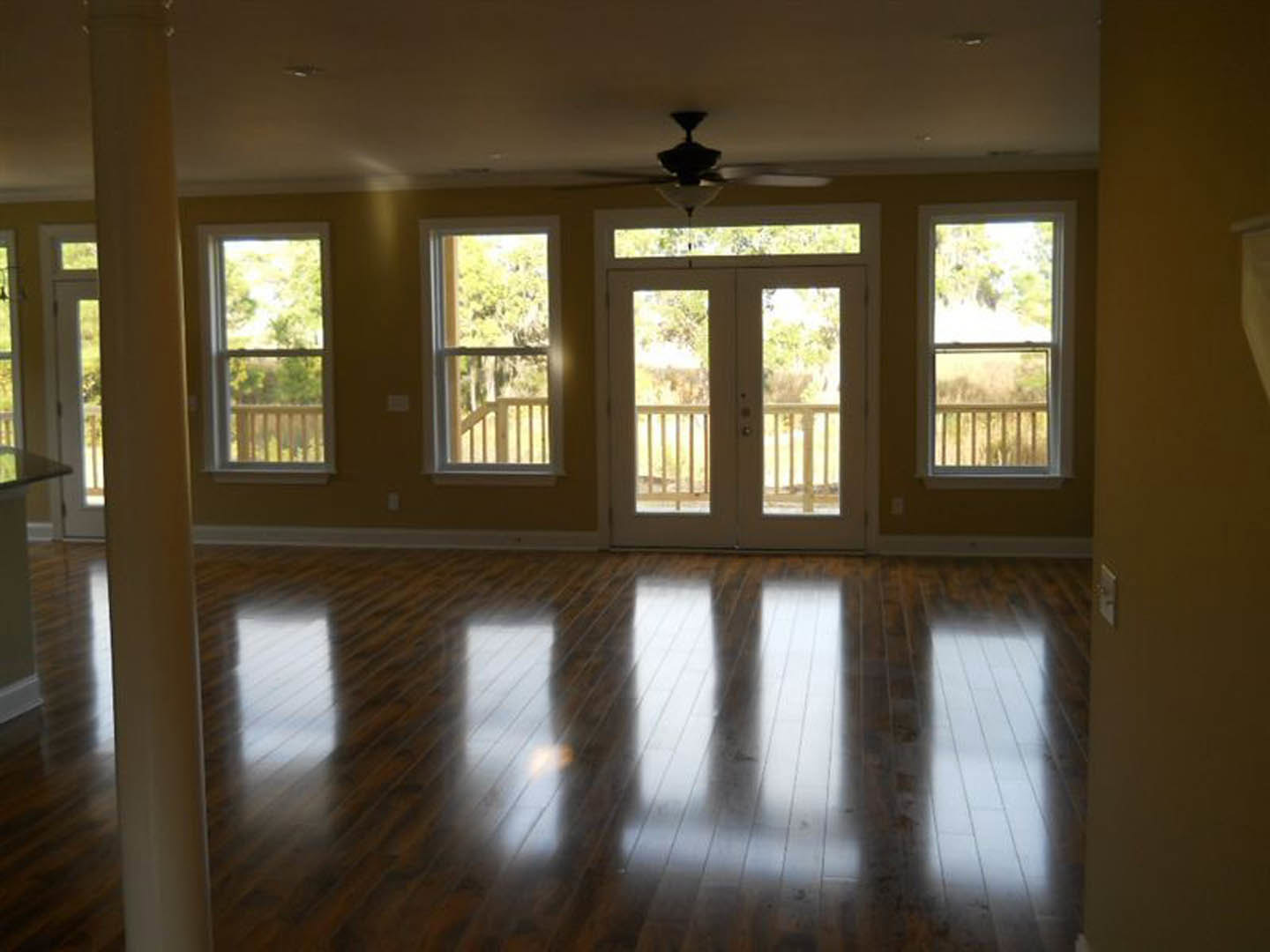 Wood floor room with ceiling fan, glass-paneled double doors, pillar detail, two windows overlooking trees, balcony access, and daylight reflecting on polished flooring