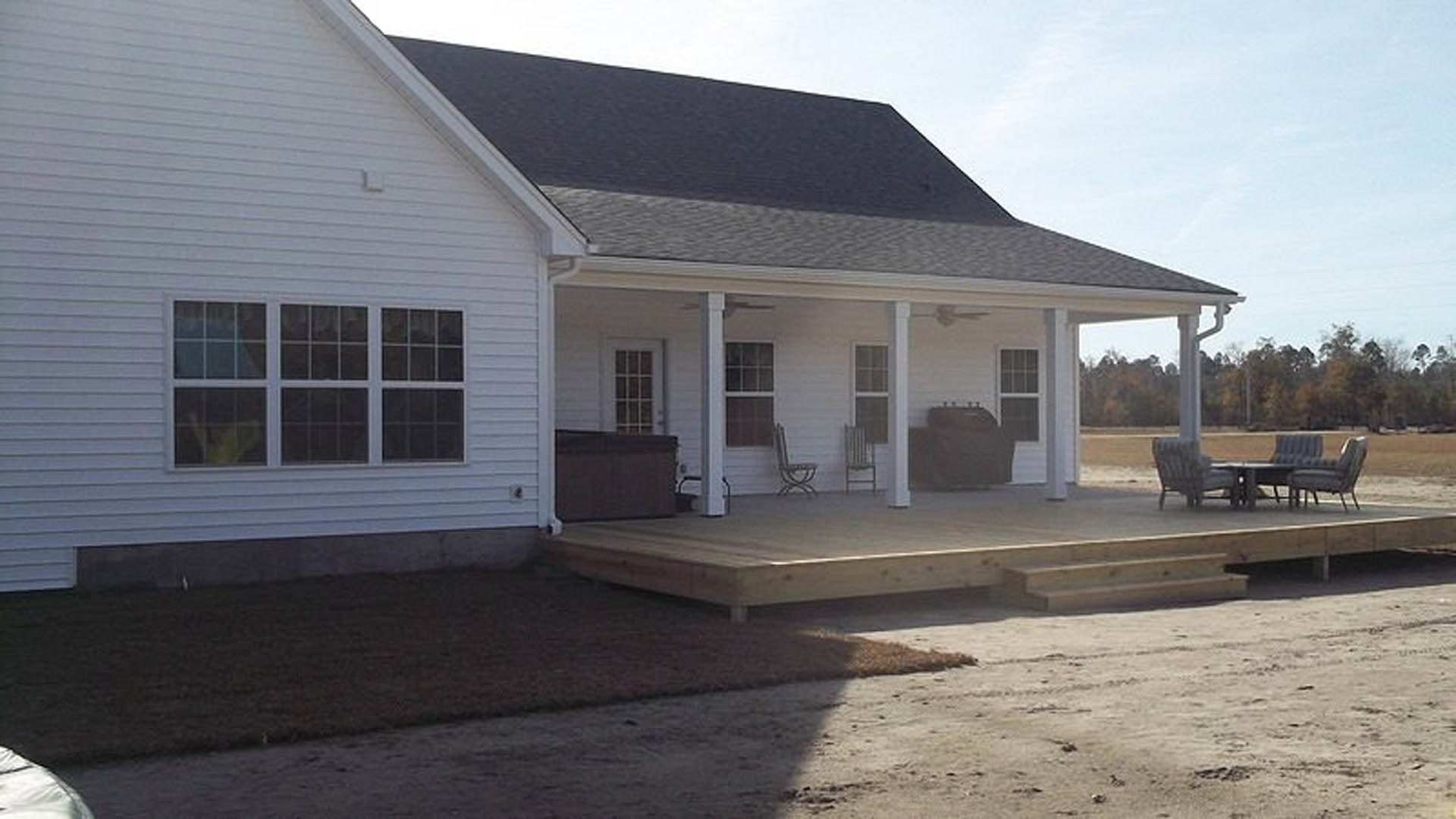 White cottage-style home with horizontal siding, covered porch featuring wooden deck and steps, metal-framed chair and small table near a large window, blue sky in background.