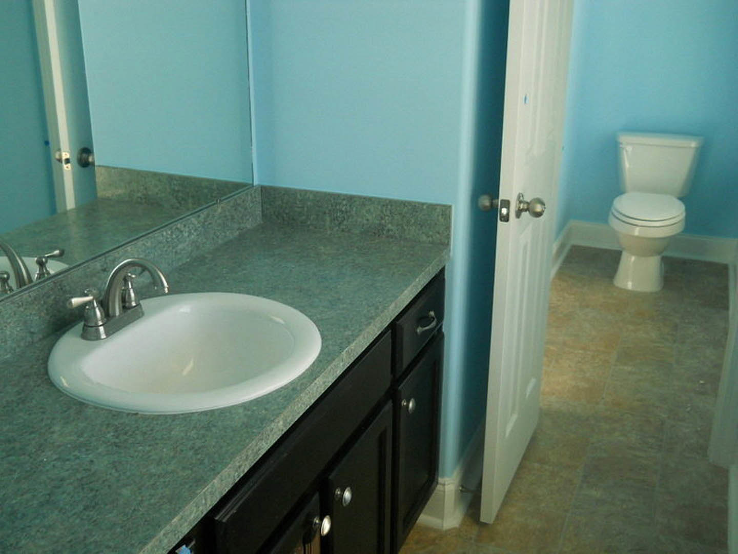 White porcelain sink set in a stone countertop beside a modern toilet, chrome faucet, tiled walls, and a brushed metal door knob visible in foreground