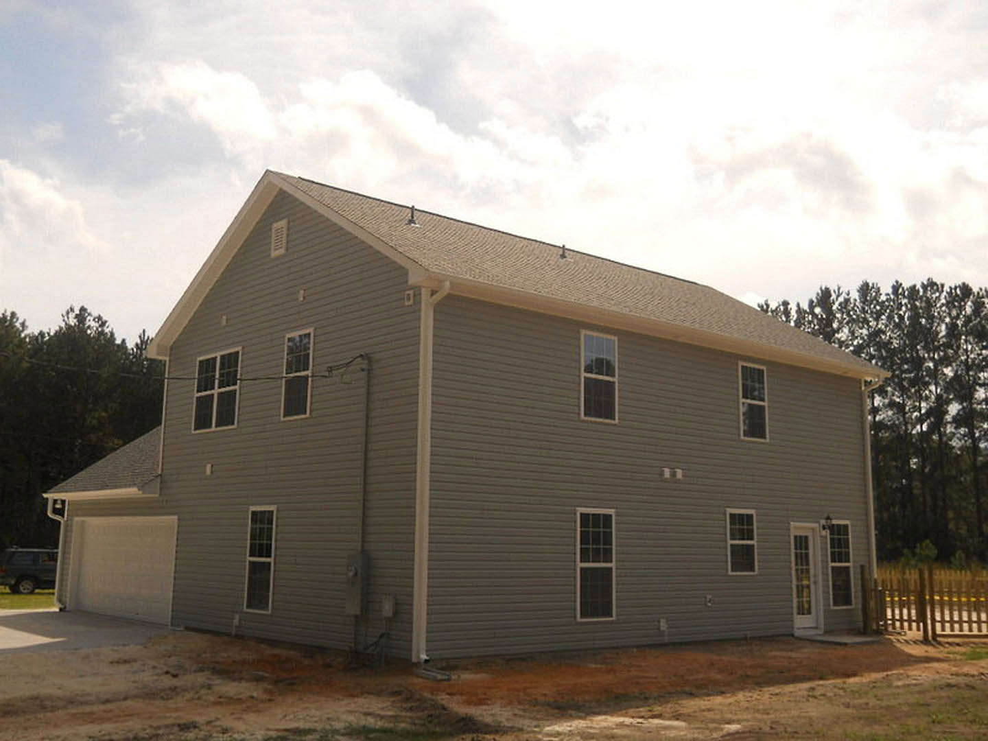 Grey house with white trim under construction, multi-pane windows, partial fence with poles, car and man in parking lot, Rockingham Meeting House visible in background, cloudy sky