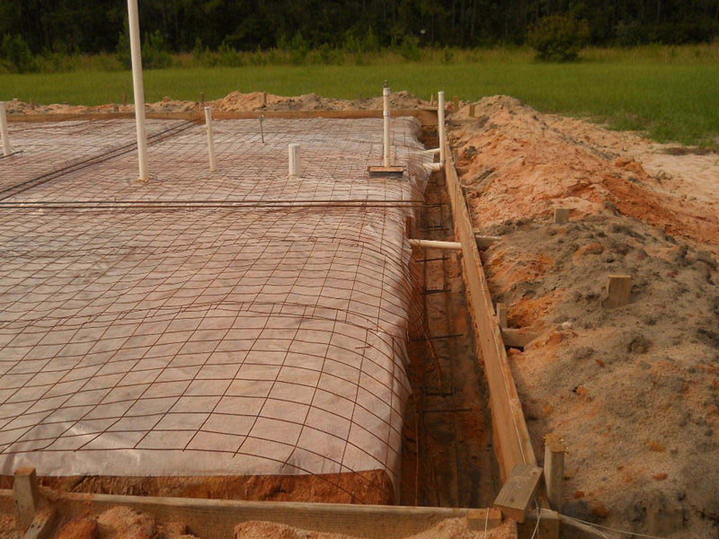 Concrete foundation under construction with wire mesh reinforcement, white pole in foreground, pile of sand and dirt nearby, grassy area and trees in background