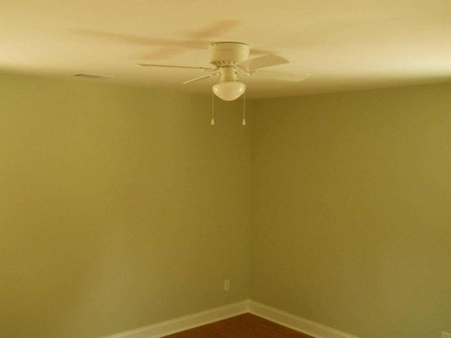 Ceiling fan with integrated light fixture mounted on white plaster ceiling above wood floor and white baseboard in corner of residential room
