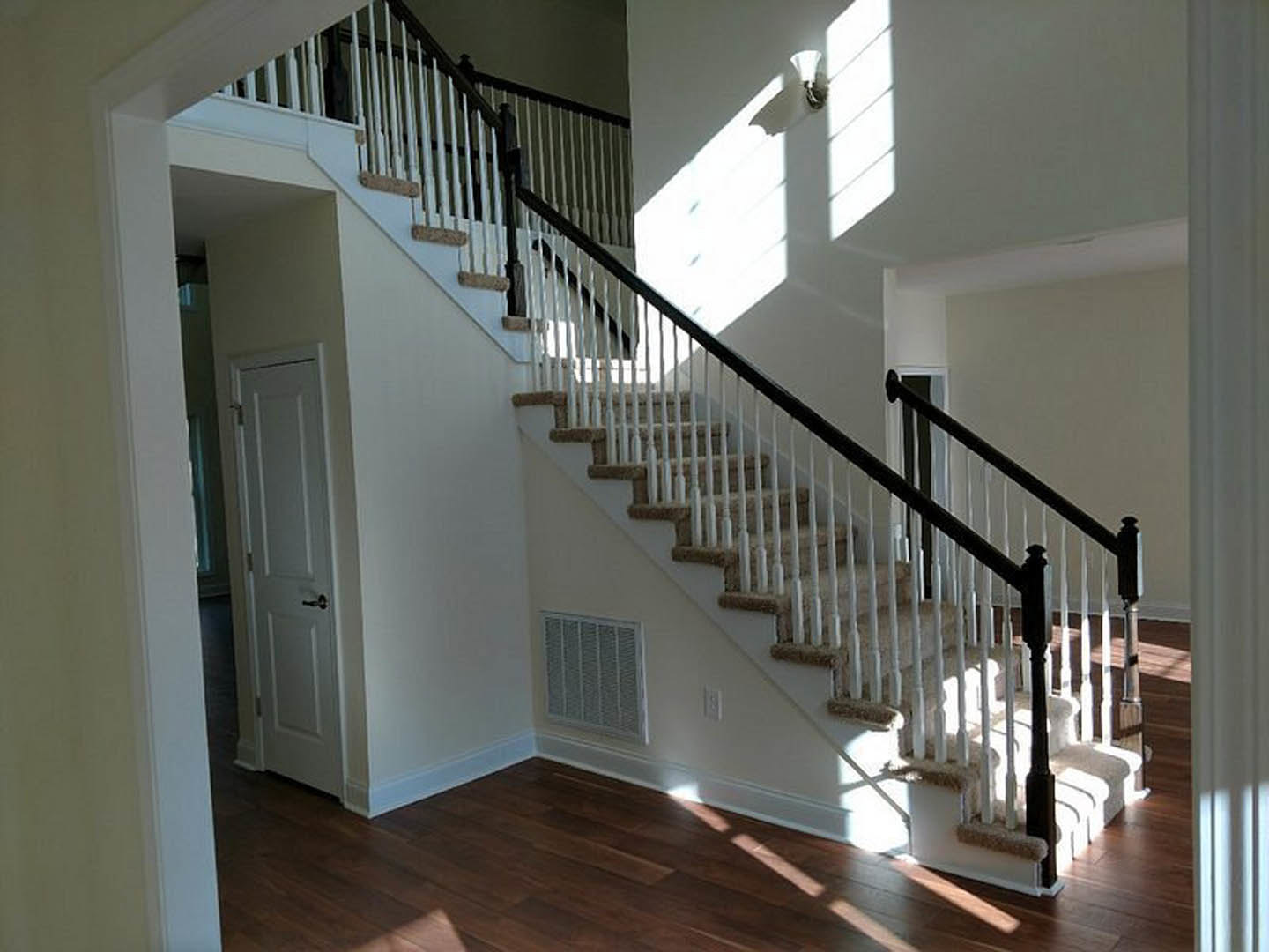 White carpeted staircase with wooden handrail, white door with silver handle, vent on wall, sunlight illuminating hardwood floor, shadow of a cat cast on white wall.