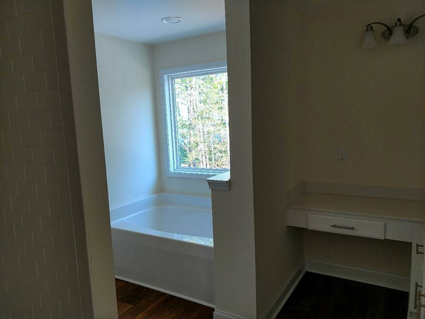 Freestanding bathtub beneath large window with tree views, white shelf and drawer, modern light fixture, neutral walls and flooring