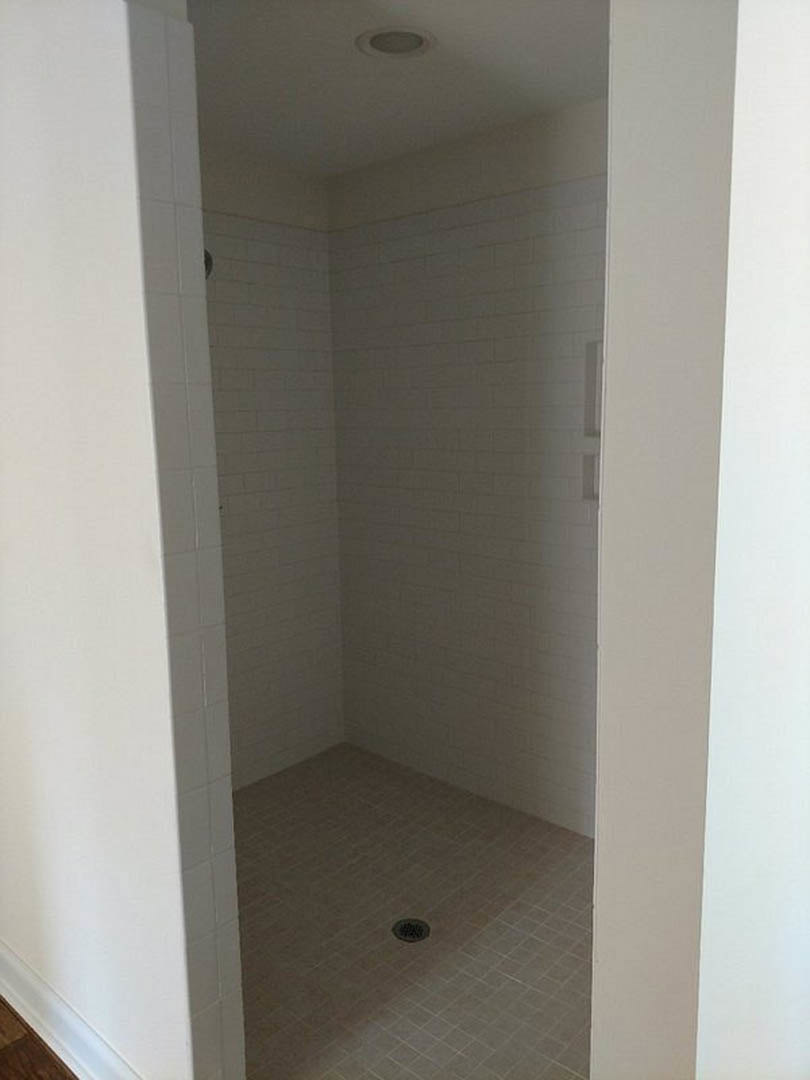 Walk-in shower featuring white tile walls, tile floor with central drain, and open doorway; adjacent white plaster walls and wood flooring visible in bathroom interior.