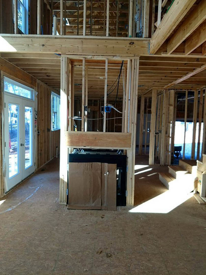 Exposed wood framing with ceiling beams, electrical wiring, and insulation visible; brown paper bag sits on unfinished black fireplace; construction materials scattered on