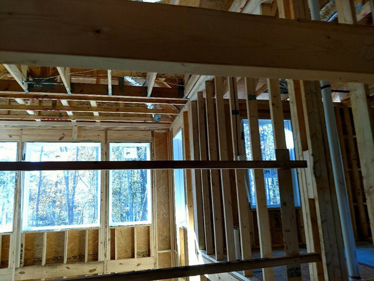 Exposed wood framing and beams in a partially constructed room with large windows, hardwood planks, and visible ceiling and wall structure.