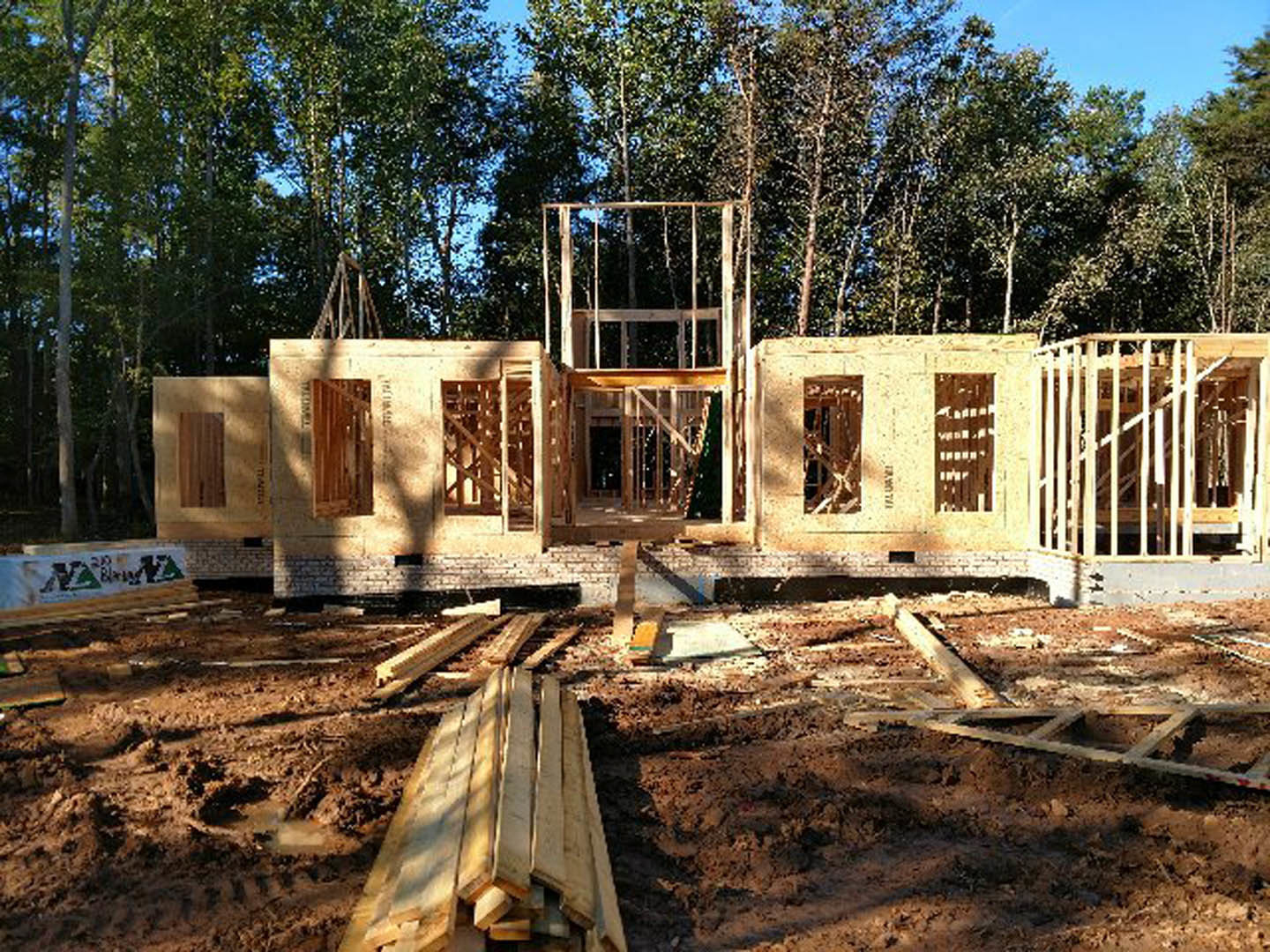 Framed wooden house under construction with exposed beams, pile of lumber on dirt ground, leafy trees and blue sky in background