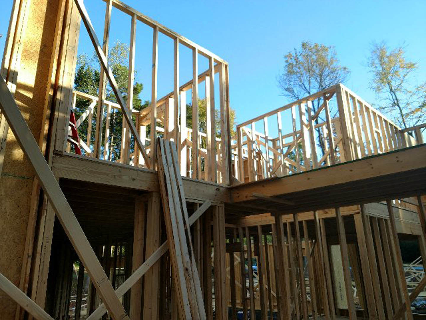 Wooden house frame under construction with exposed beams, partially built stairs, and surrounding trees and fencing under a clear sky