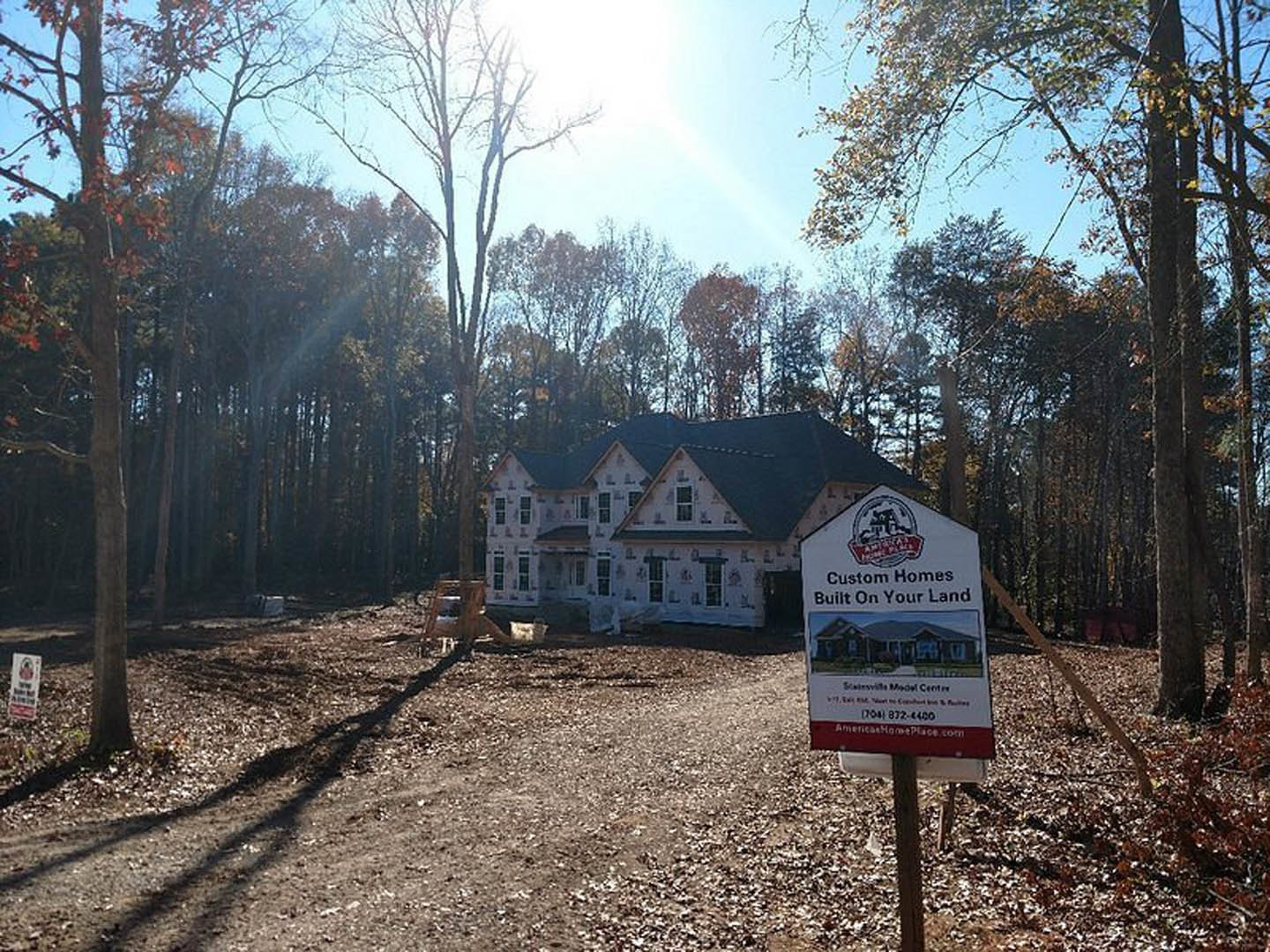 Two-story house with black roof, surrounded by trees and blue sky, sign posted in center of grassy yard, dirt path leading to entrance, parked car partially visible