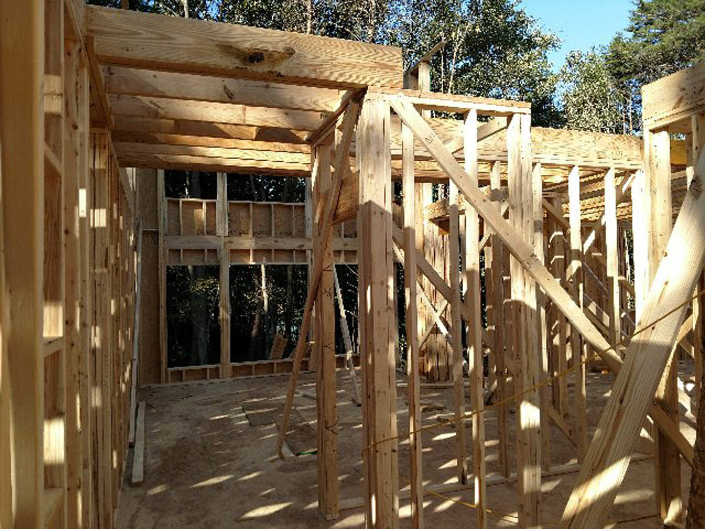 Wooden framing structure of a house under construction with exposed beams and planks, surrounded by dirt ground, trees, and a fence under a clear sky