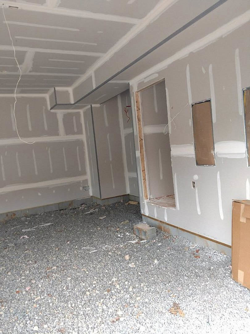Open room with white plaster walls, large windows, wooden door, brown pillar with white trim, light-colored flooring, and exposed brick accents