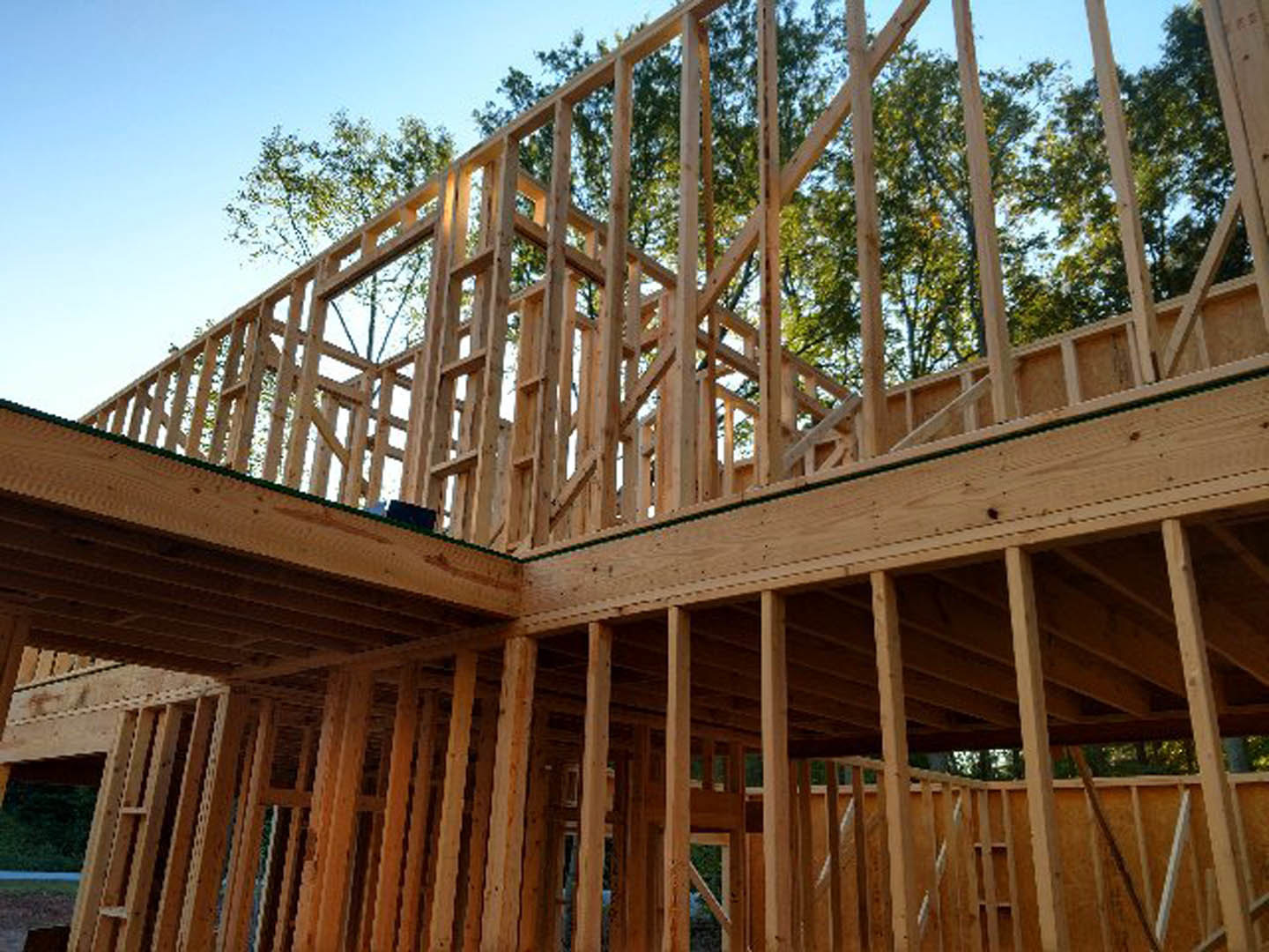 Wooden house frame under construction with exposed beams and lumber, surrounded by trees and a fence under a clear sky