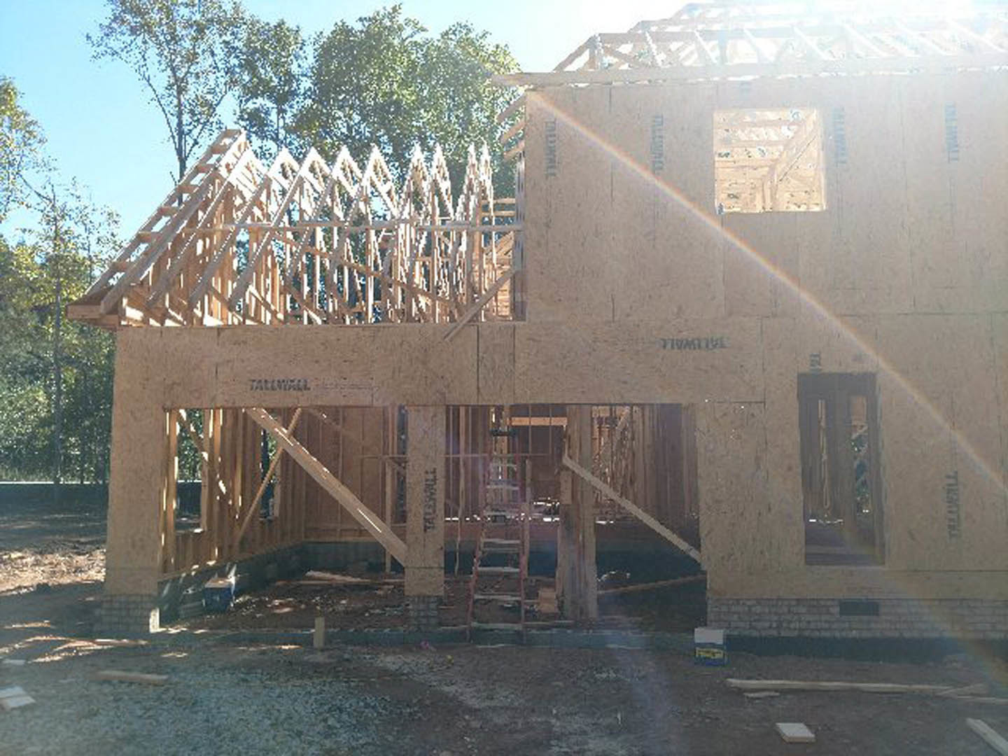 Wooden house frame under construction with exposed beams, ladder leaning against structure, trees and sky in background