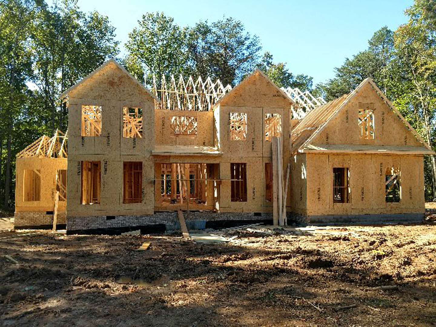 Wood-framed house under construction with exposed beams, unfinished exterior walls, and surrounding mature trees in the background