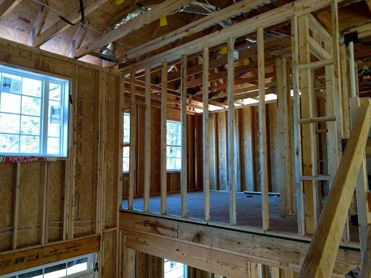 Exposed wooden framing and beams inside a house under construction, window with a sign visible, unfinished walls and insulation
