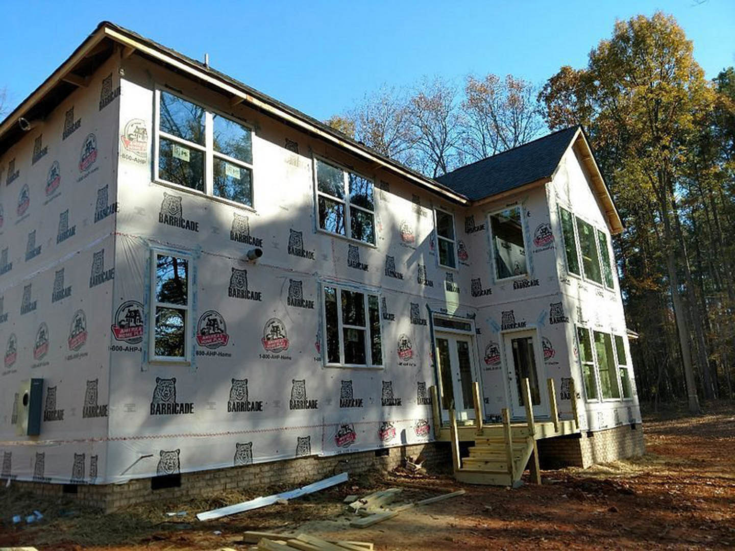 Partially built house with exposed framing, plastic sheeting covering exterior walls, several installed sash windows, and trees visible in the background